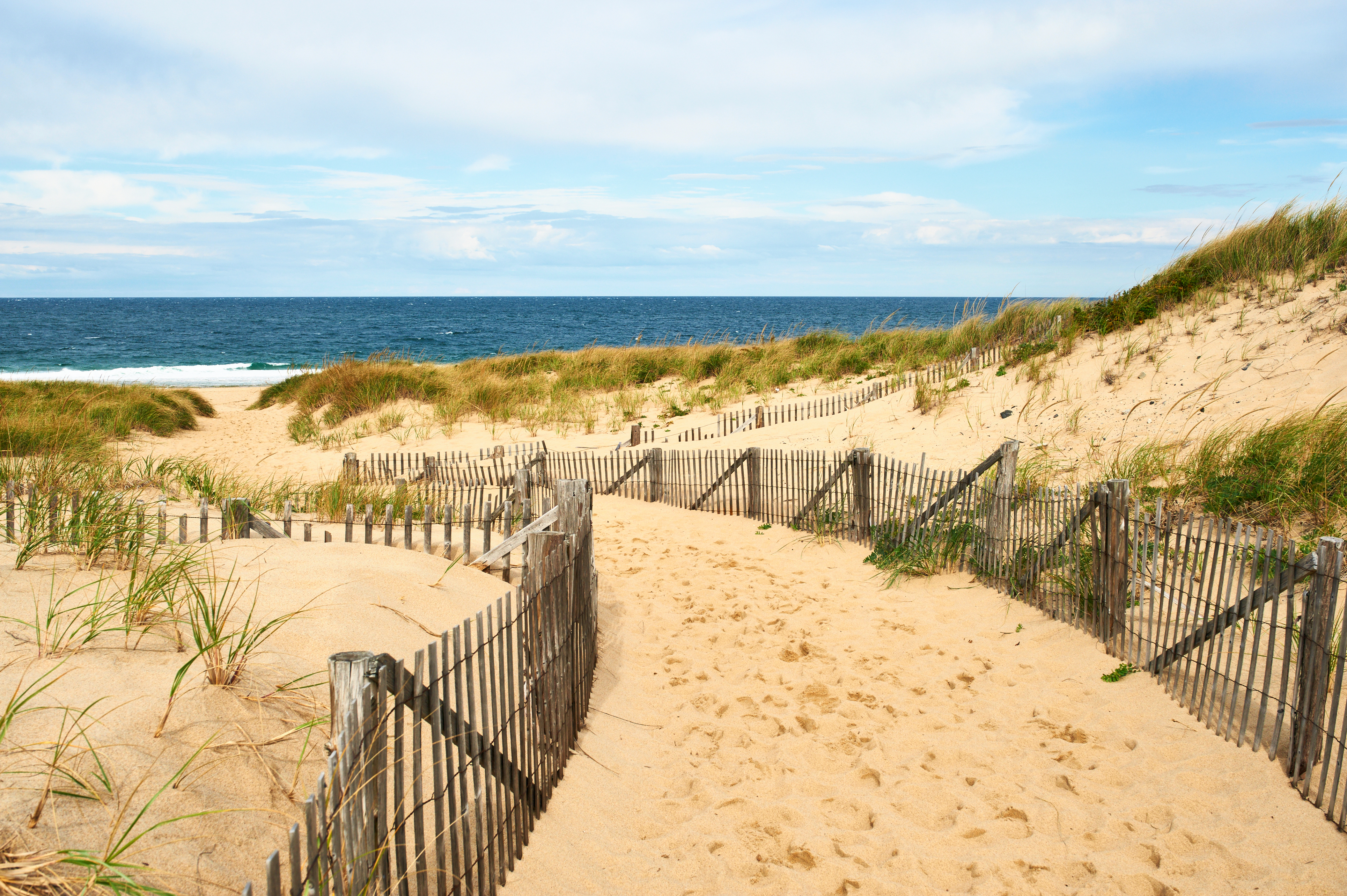Sandy beach path lined with wooden fences and grass-covered dunes leading to the ocean under a partly cloudy sky.