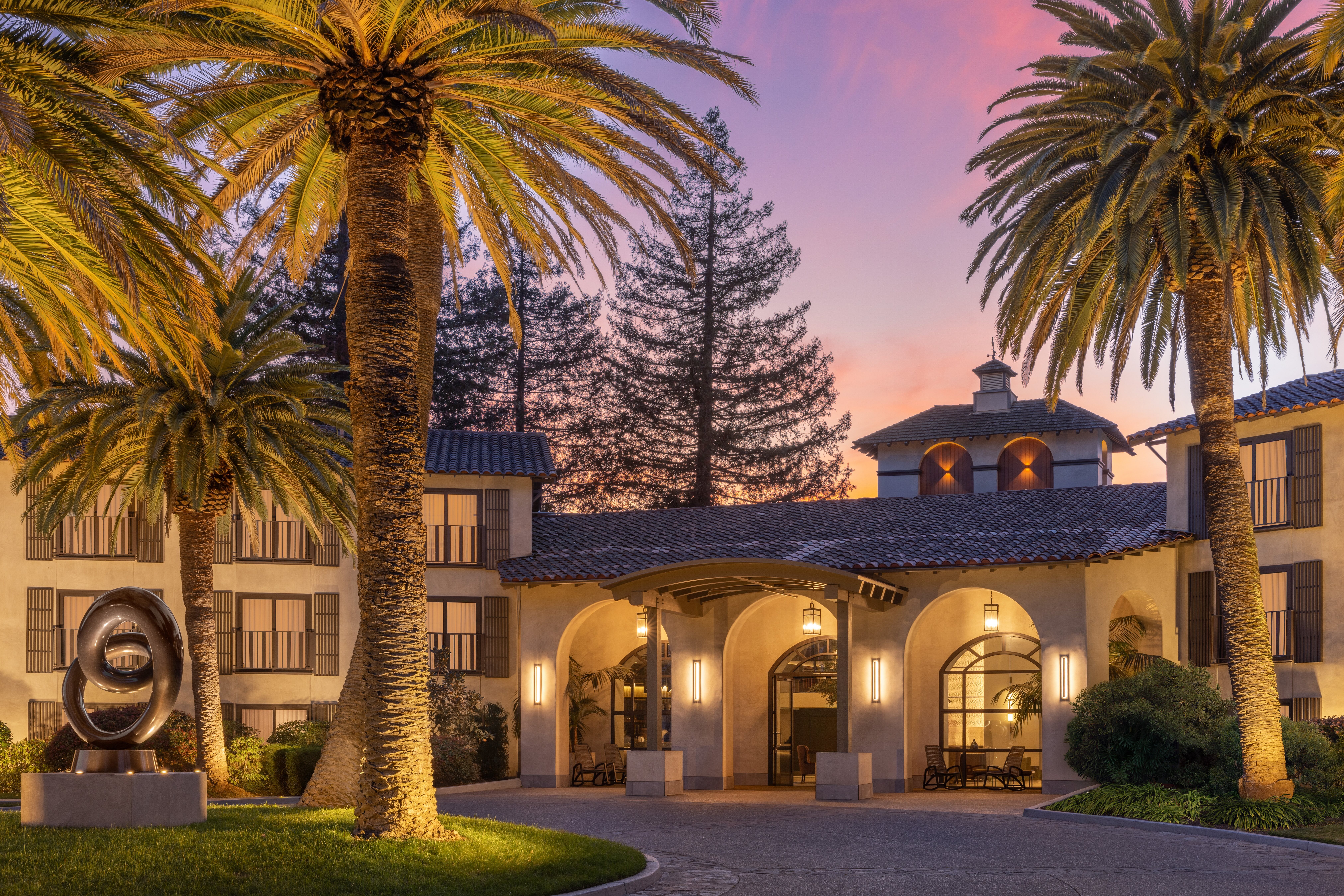 Elegant entrance to Casa Mani Resort Napa Valley, Curio Collection by Hilton with arched doorways, palm trees, and a modern sculpture at sunset.