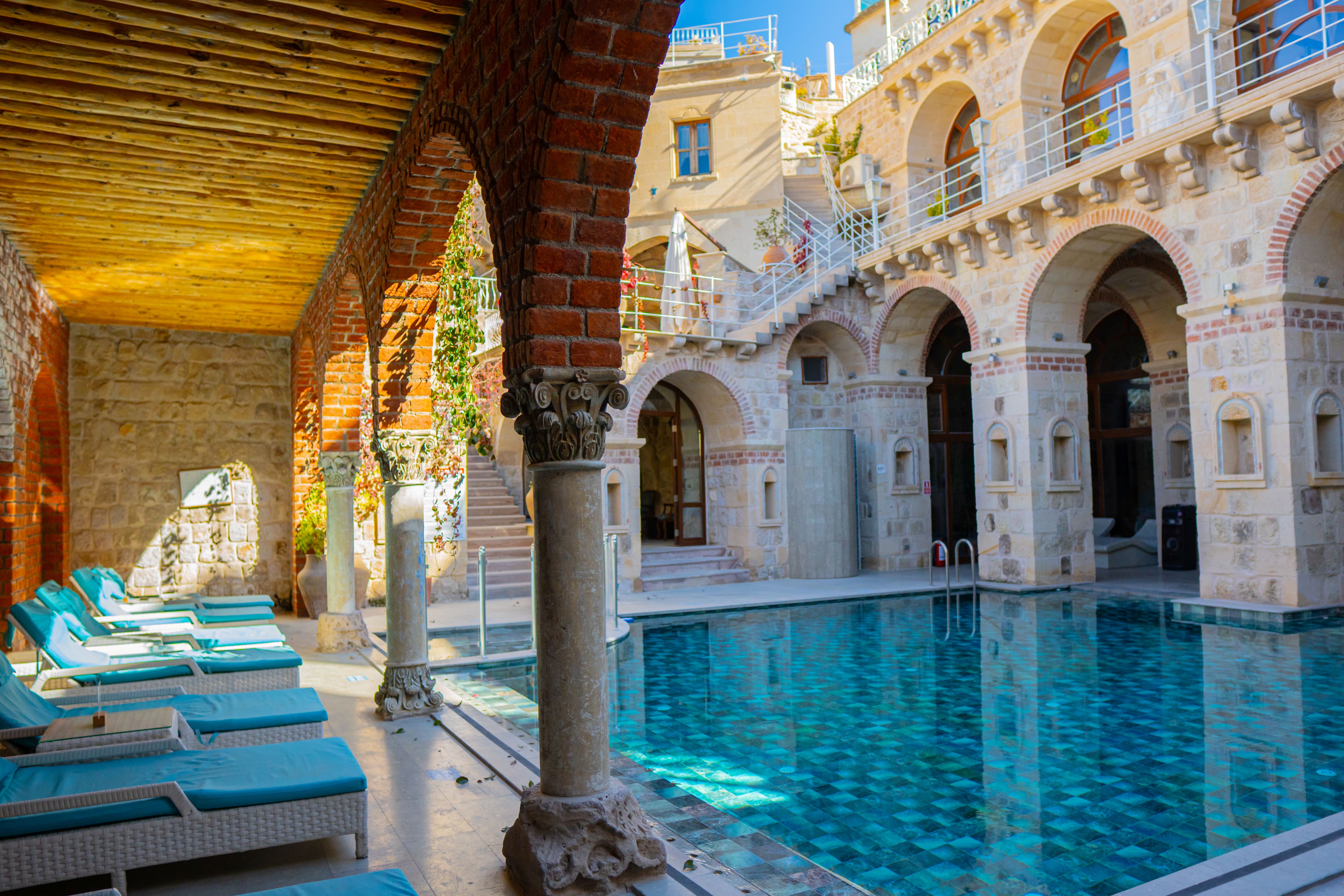 Outdoor pool with turquoise water surrounded by stone arches and a lounge area with blue cushioned chairs under a wooden ceiling with brick arches.