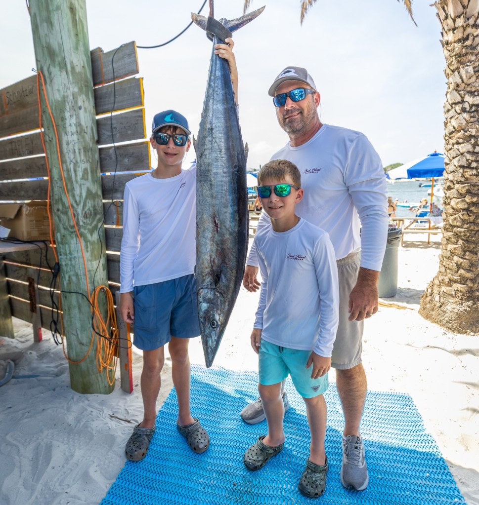 A dad and two kids wearing sunglasses and casual beachwear stand on a blue mat in the sand, holding a large fish hanging vertically.