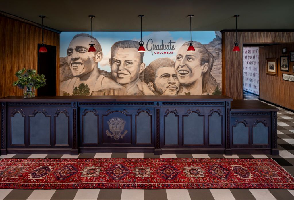 Reception area with a dark wooden desk, red pendant lights, a colorful patterned rug, and a mural of four smiling faces with "Graduate Columbus" text above.