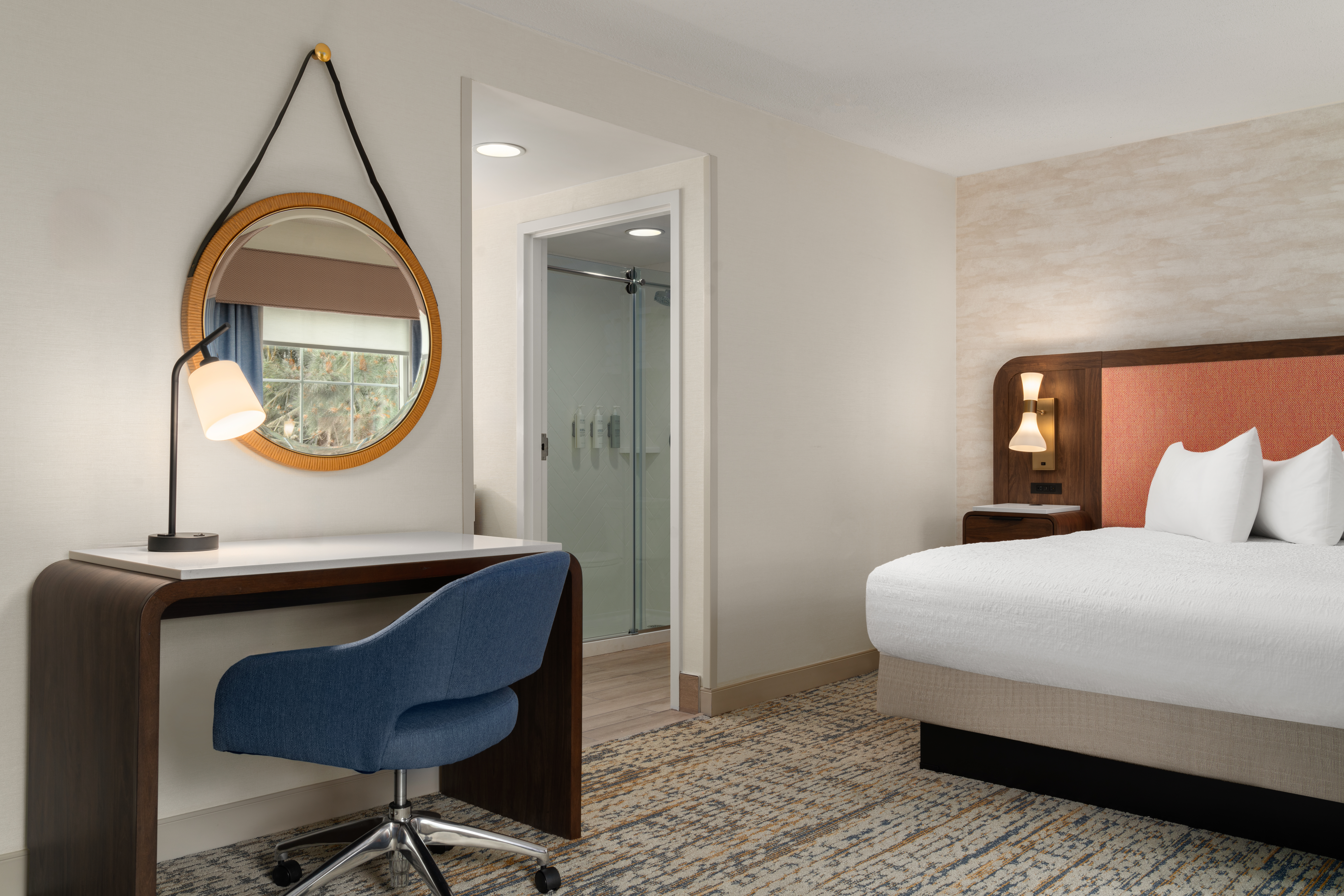 Modern hotel room with a bed, bedside lamp, desk with a lamp, blue chair, round mirror, and a bathroom with a glass shower door.