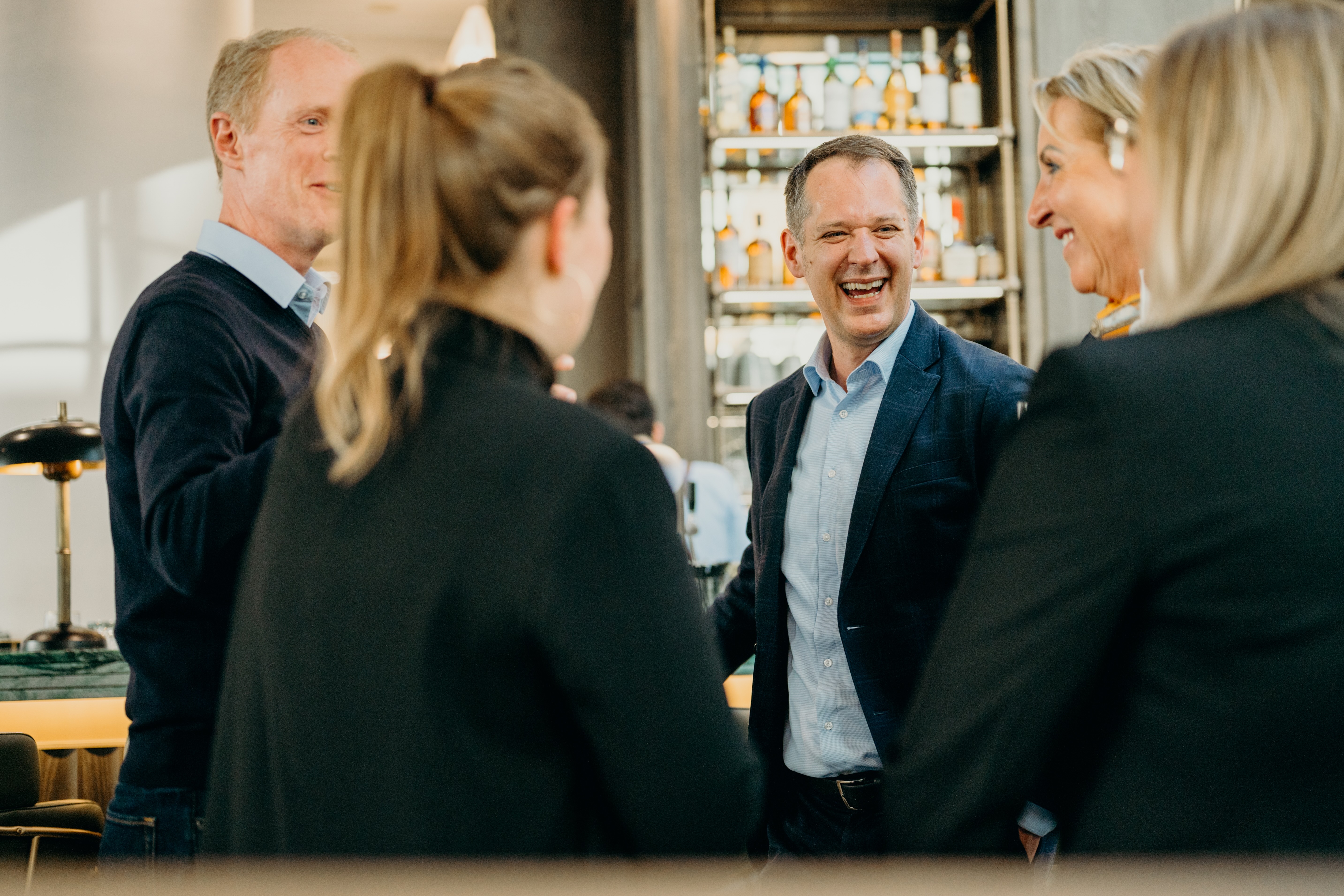 Five people in business attire are engaged in a lively conversation in a well-lit room with shelves of bottles in the background.