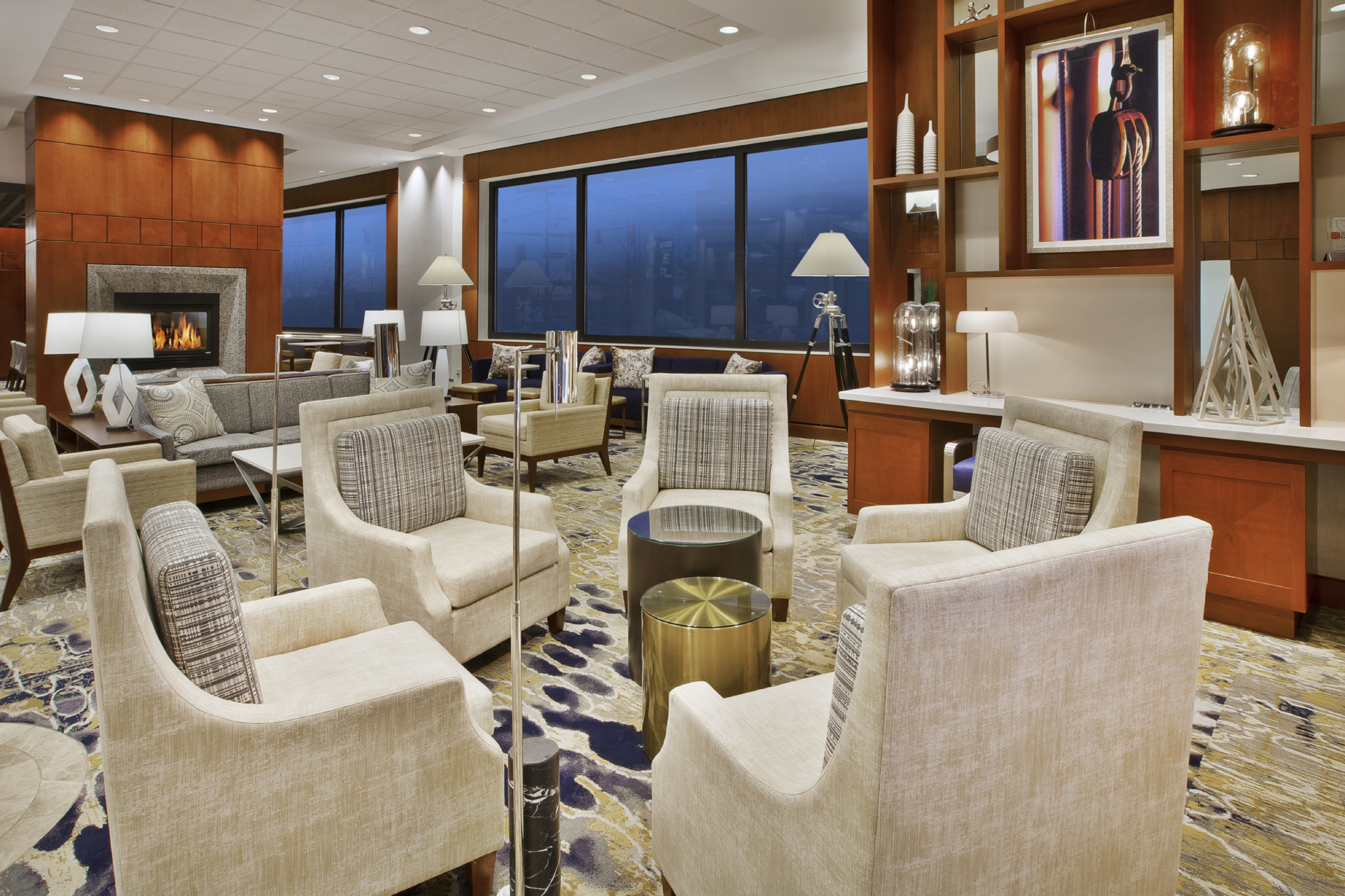 Modern lounge area with beige armchairs, patterned cushions, a fireplace, and large windows with a nighttime view.