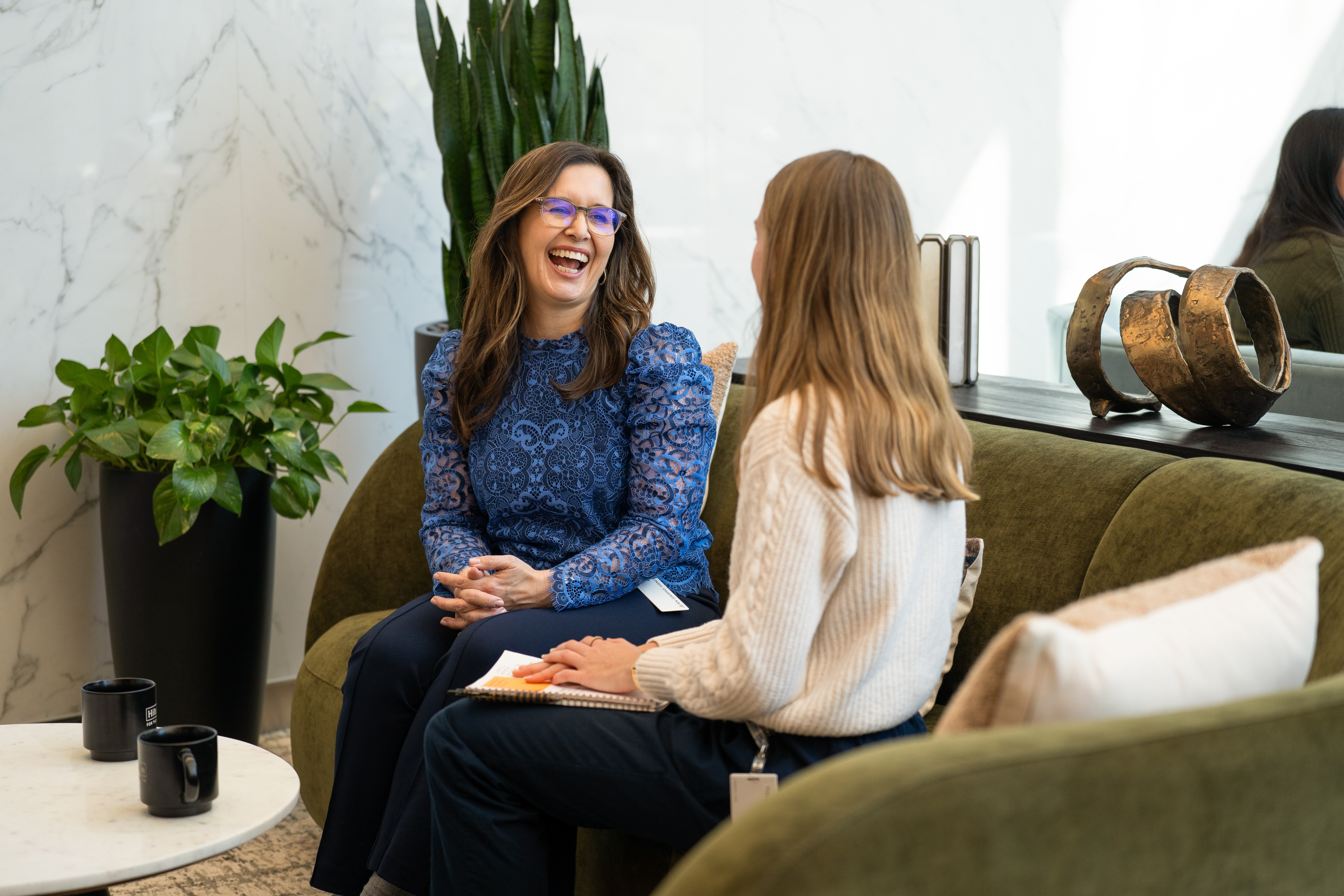 Two women sitting on a green couch, one laughing and the other holding a notebook, in a modern, plant-decorated room.