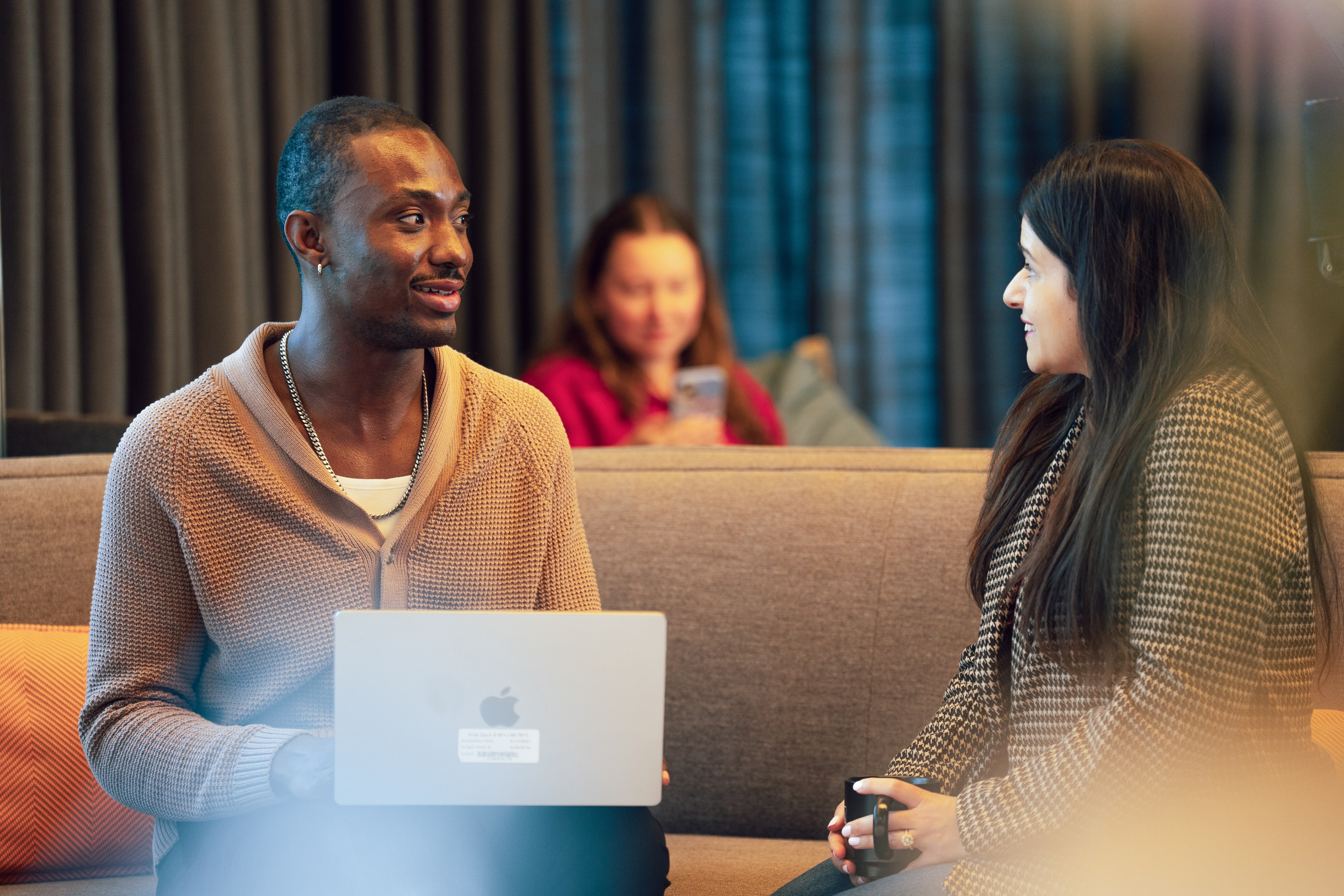 Two people sitting on a couch, one holding a laptop and the other holding a mug, engaged in conversation with a third person blurred in the background.