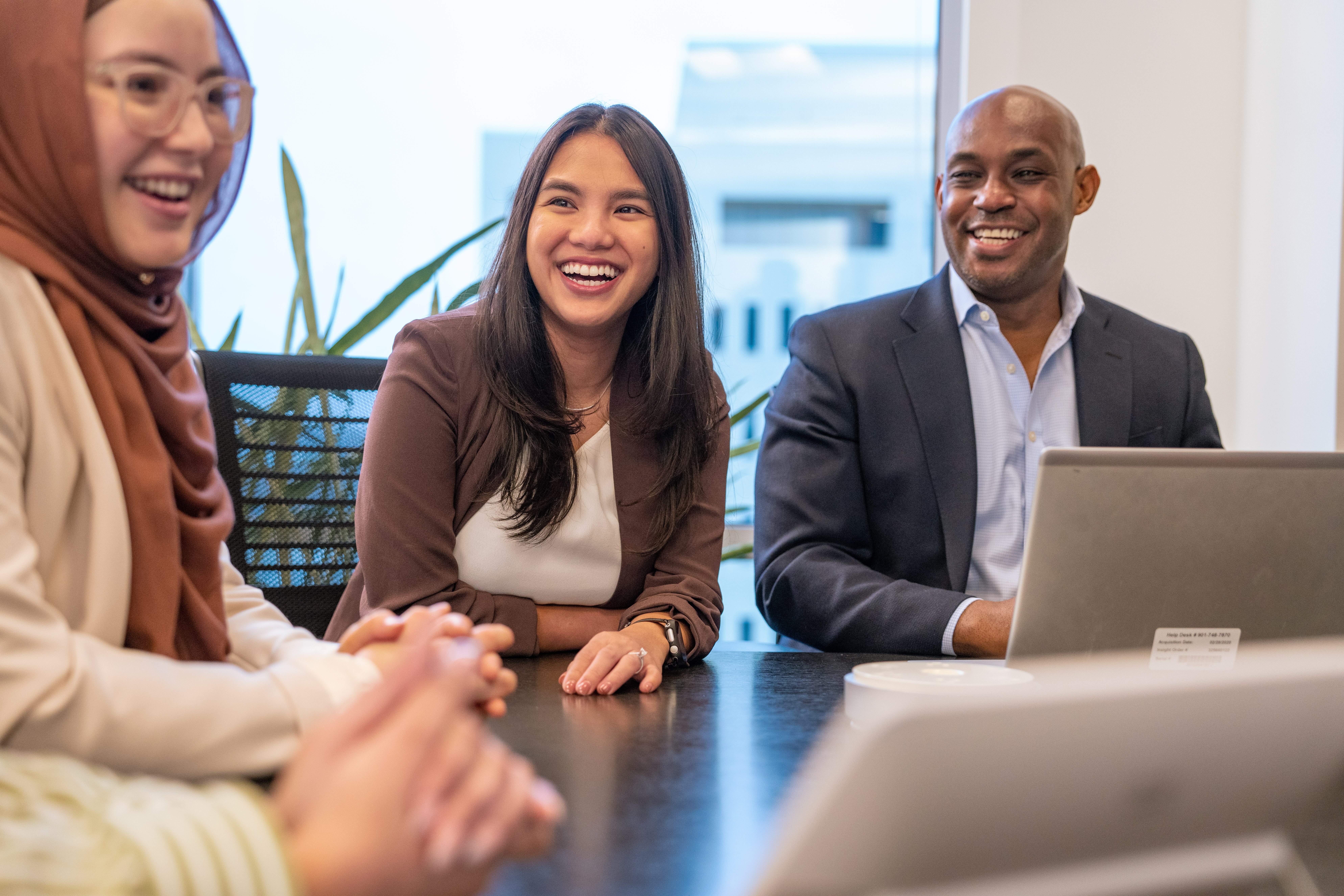 Three colleagues smiling and engaged in a meeting around a conference table with a laptop.