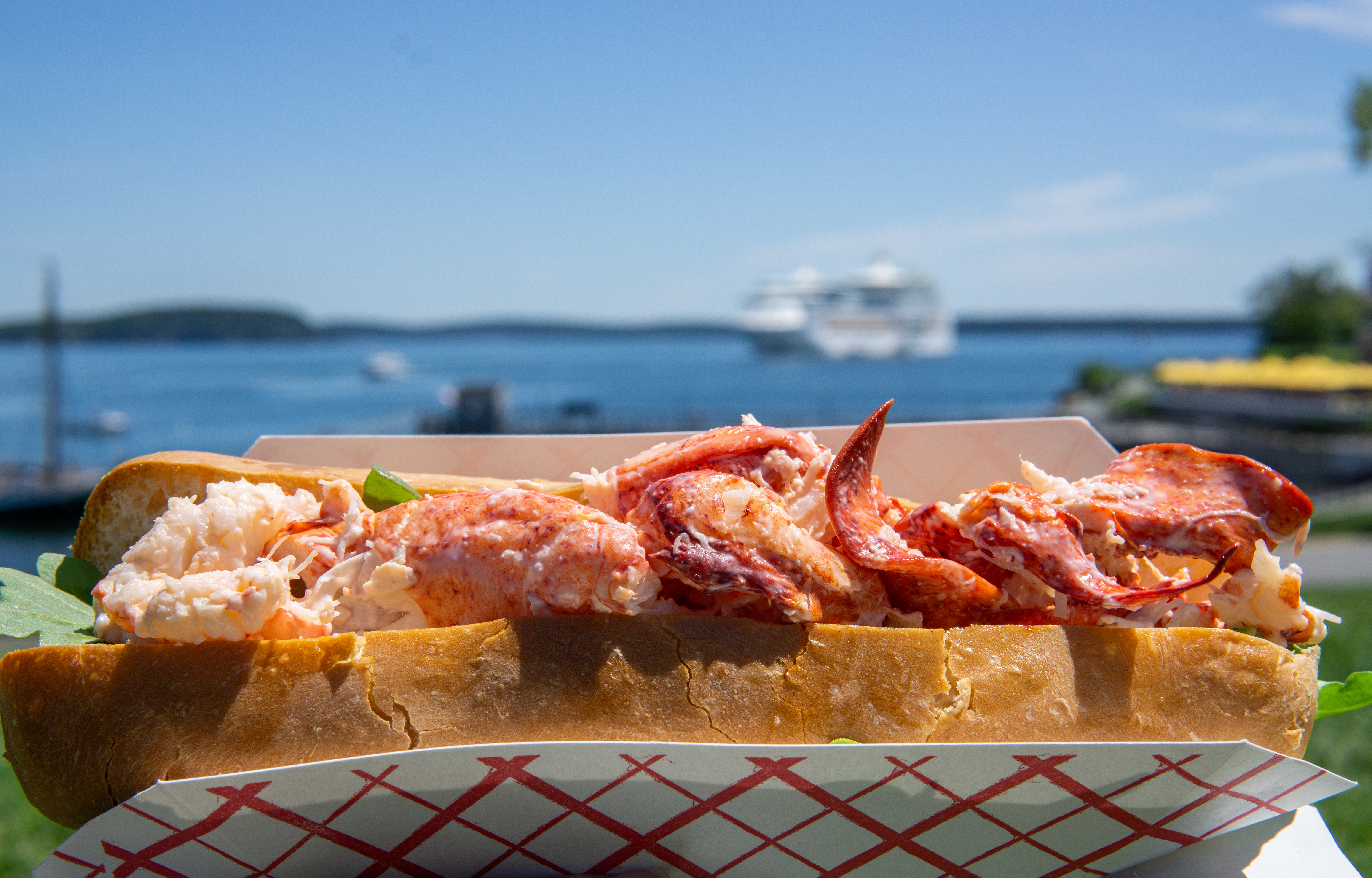Lobster roll sandwich with chunks of lobster meat in a toasted bun, held outdoors near a waterfront with a boat in the background.