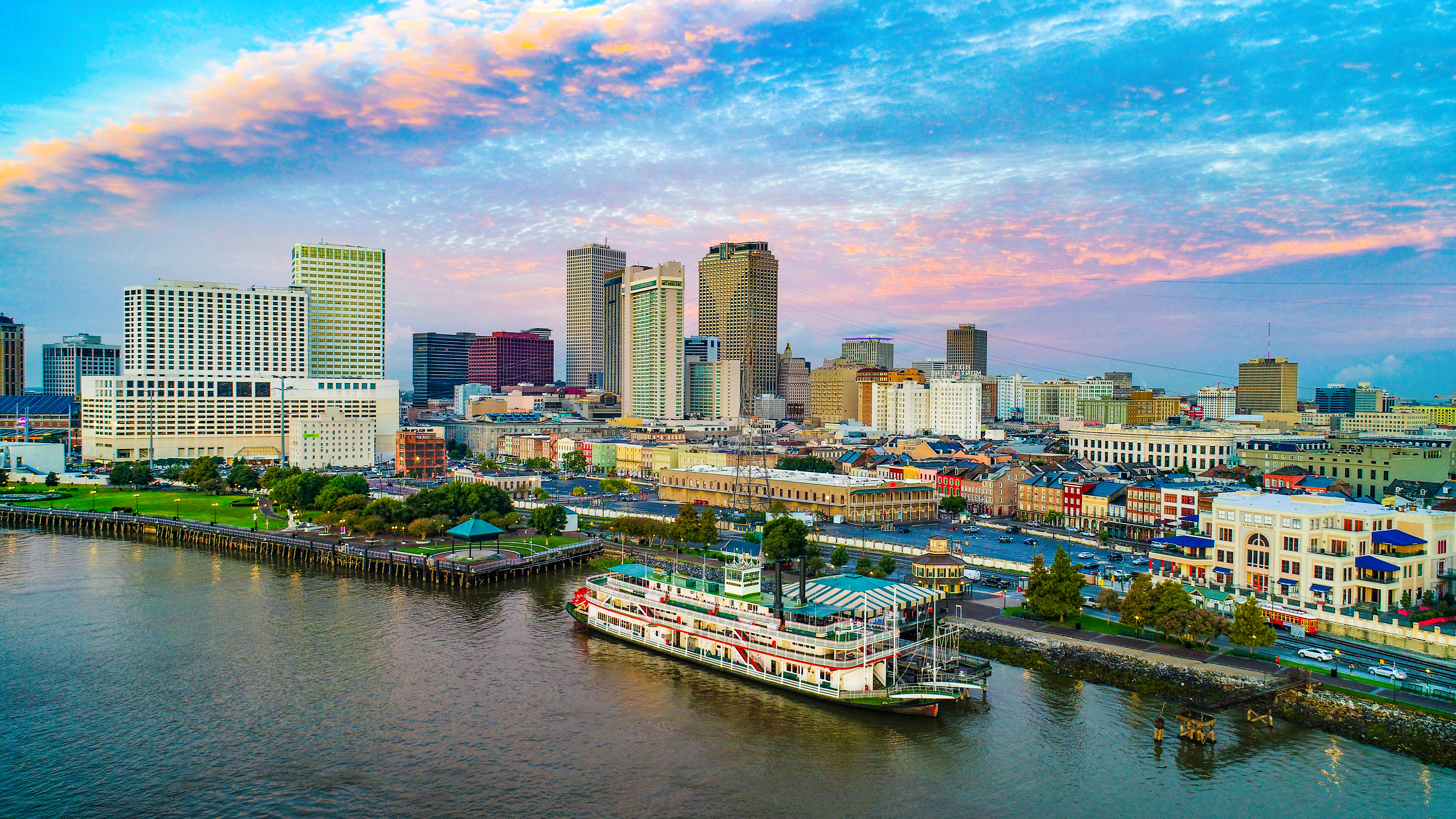 City skyline with tall buildings along a riverfront at sunset, featuring a large riverboat docked by the shore.