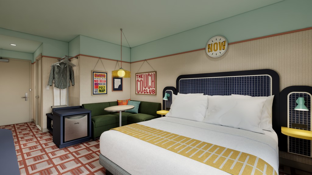 Retro-style hotel room with a large bed, green corner sofa, round table, and framed vintage posters on the wall.