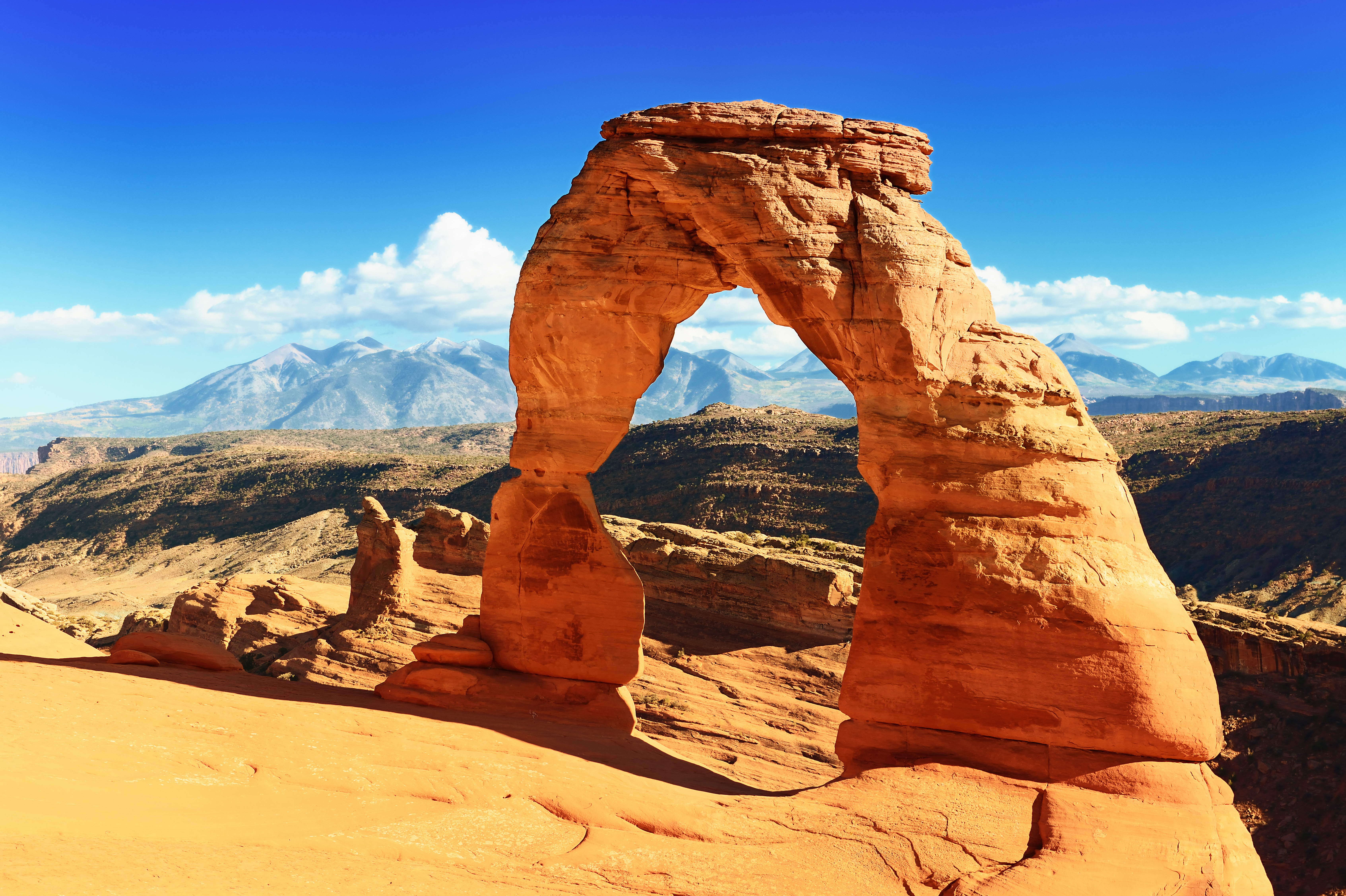 Delicate Arch, a natural sandstone arch, stands prominently against a clear blue sky with distant mountains in the background.