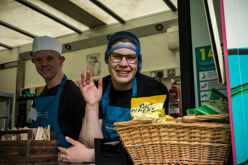 Two smiling workers in blue aprons inside a food truck, one waving and the other standing behind at Aurora Foxes