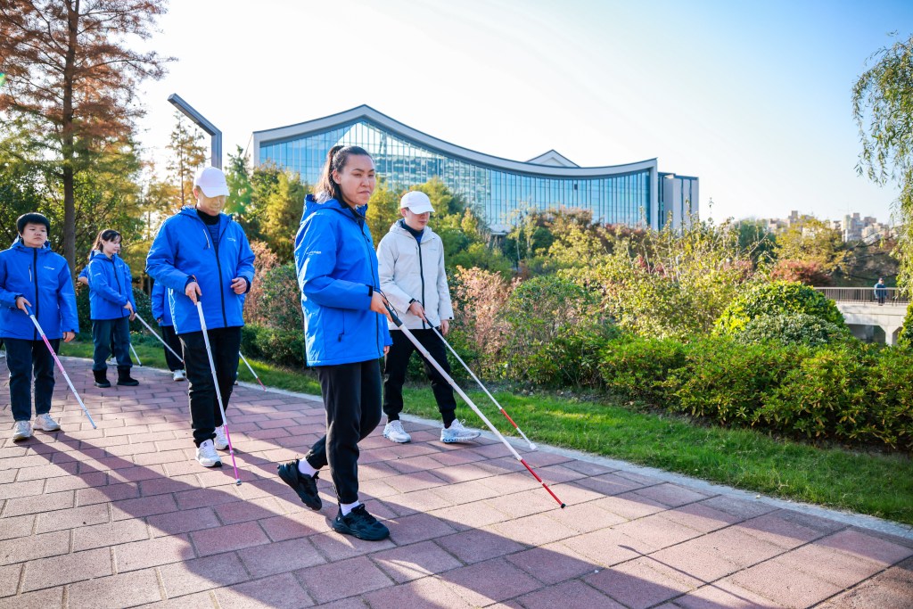 A group of people wearing blue jackets walking outdoors on a paved path, using white canes for navigation at BeYoureyeS.