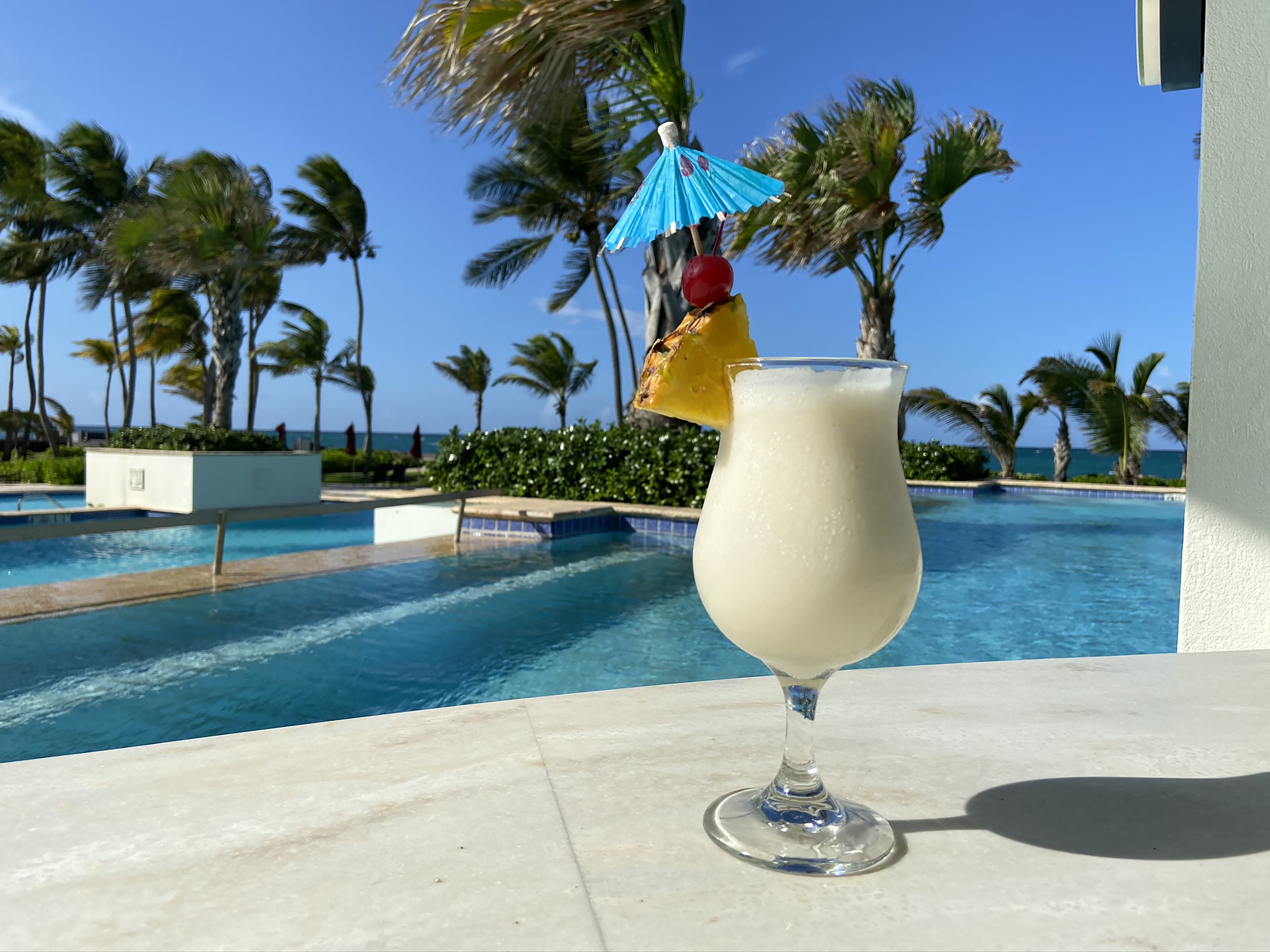 Pina Colada garnished with pineapple, cherry, and a blue umbrella by a pool with palm trees and ocean in the background.