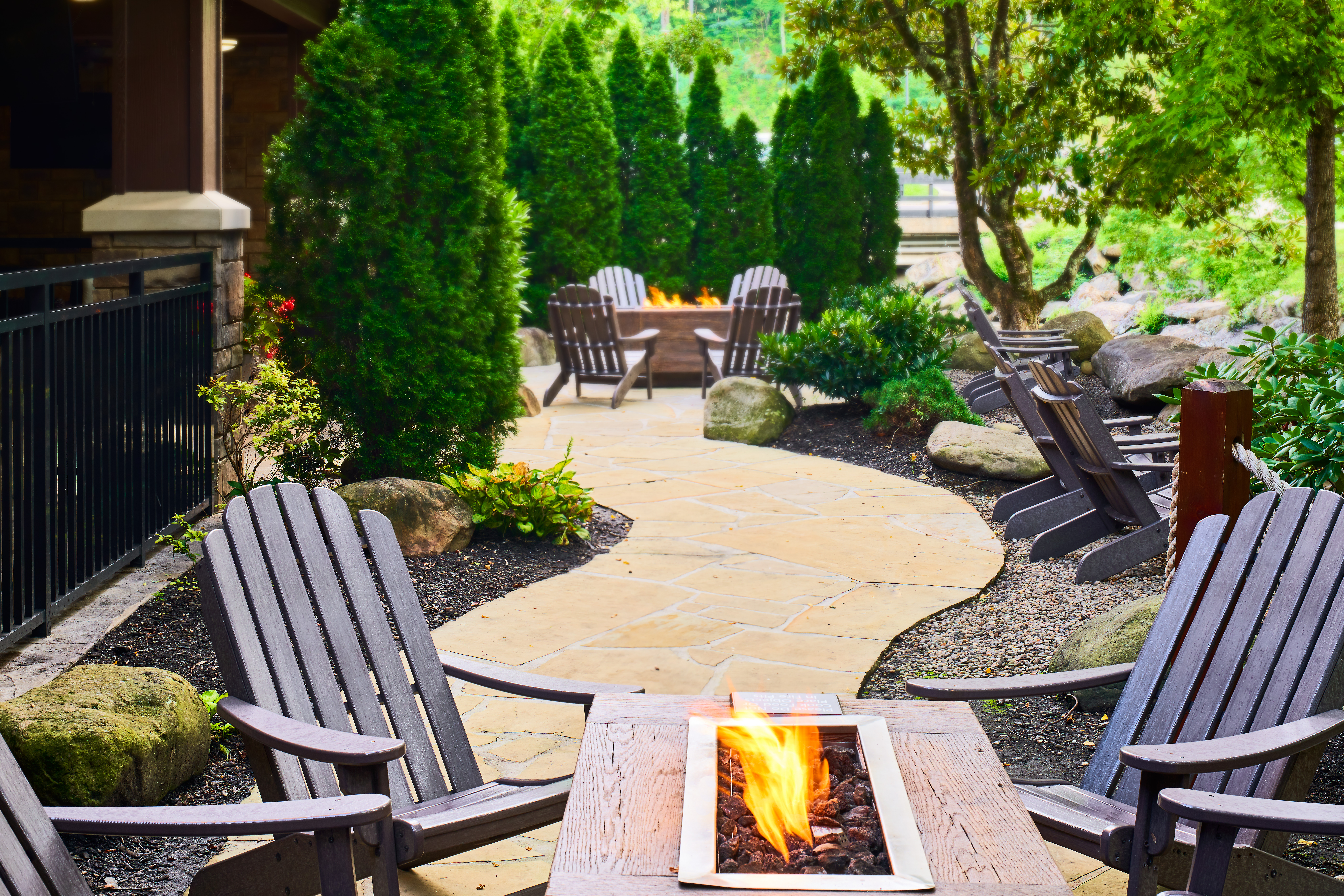 Outdoor seating area with wooden chairs arranged around fire pits along a stone pathway surrounded by greenery at the Hampton Inn Gatlinburg Historic Nature Trail.