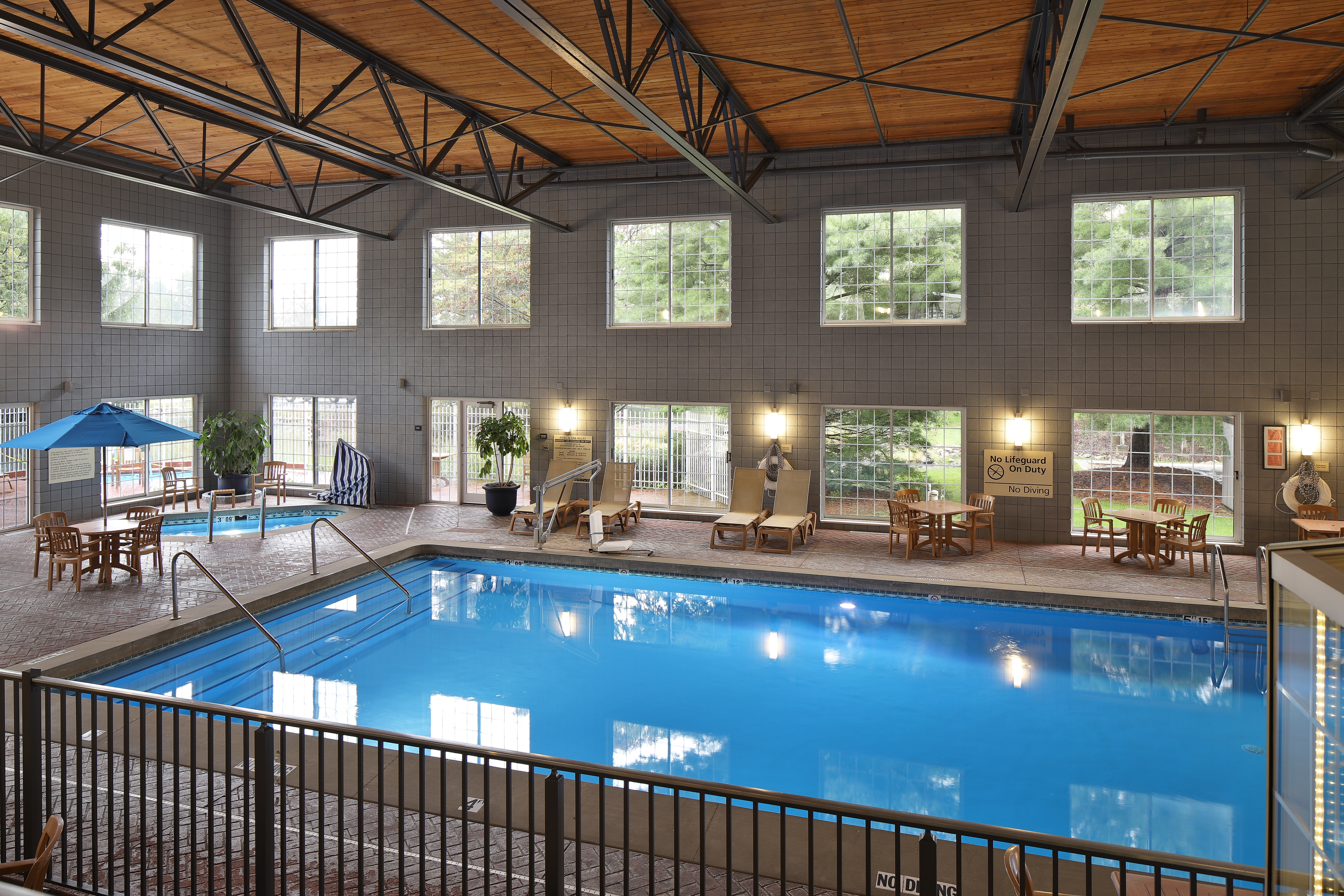 Indoor swimming pool area with lounge chairs, tables with umbrellas, large windows, and a high wooden ceiling with metal beams.