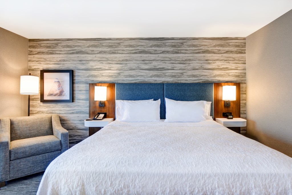 Modern hotel room with a large bed, blue upholstered headboard, two bedside lamps, a gray armchair, and abstract wall art.
