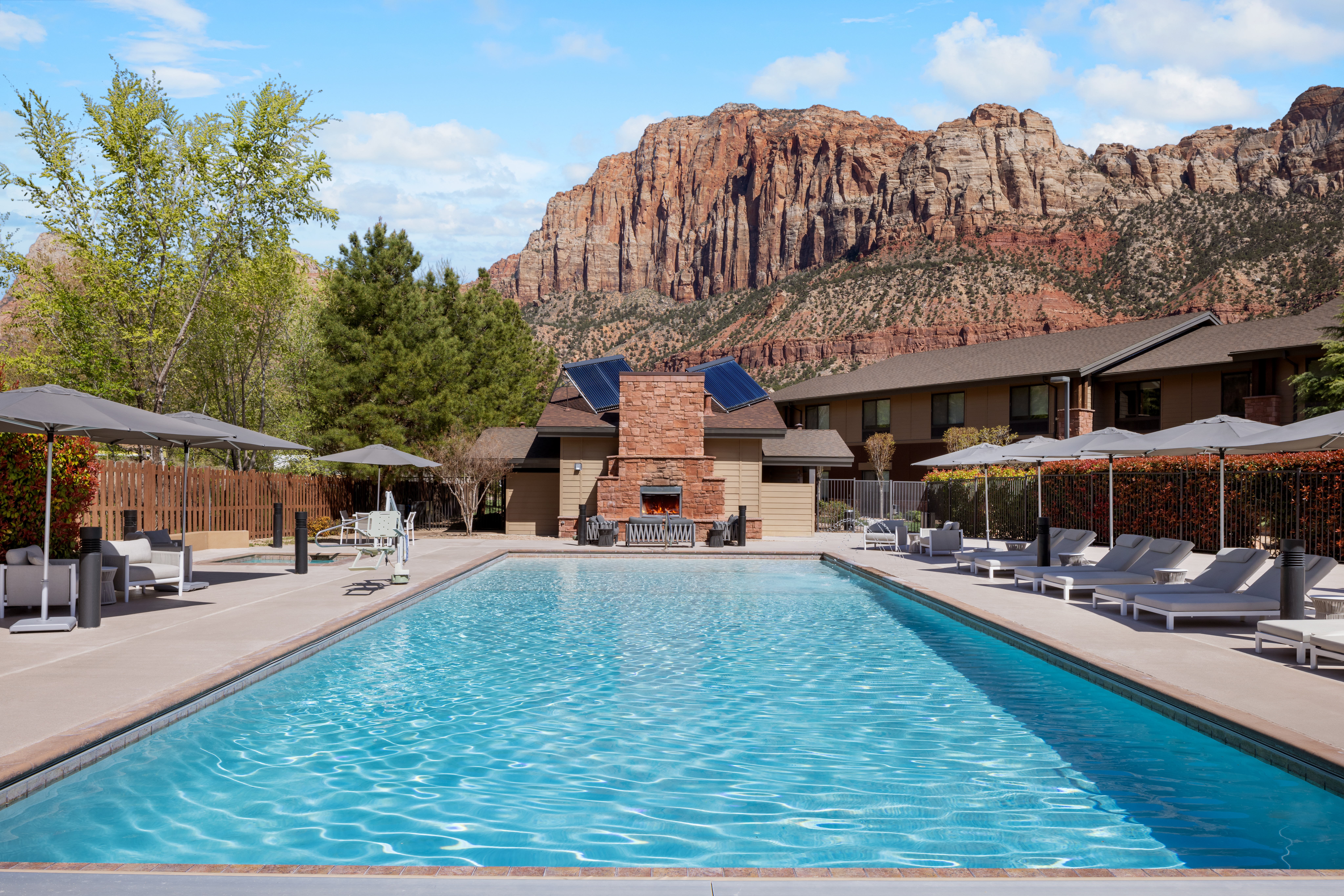 Outdoor swimming pool with lounge chairs and umbrellas, set against a backdrop of rocky mountains and clear sky.