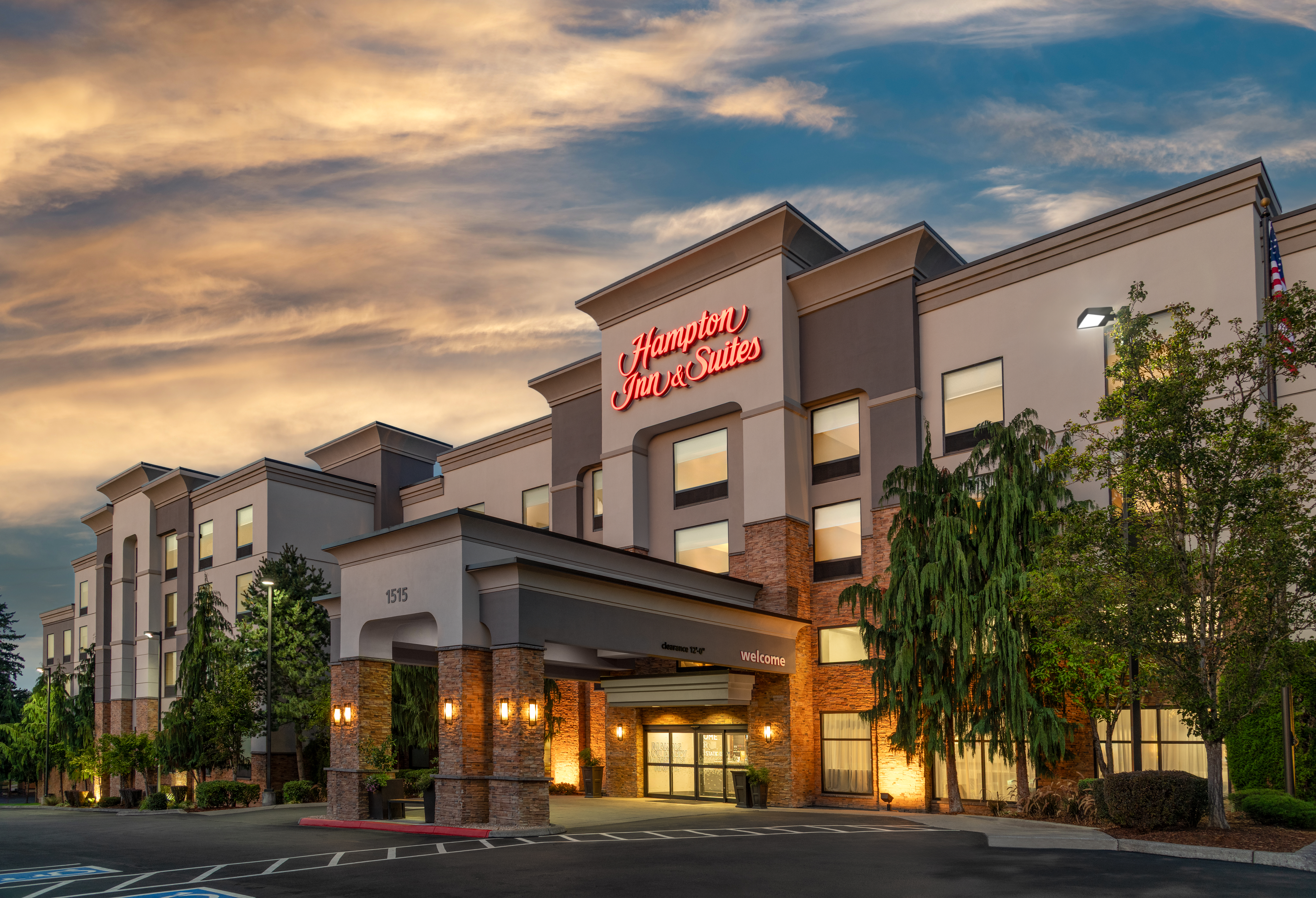 Hampton Inn &amp; Suites hotel building at dusk with illuminated entrance and partly cloudy sky.