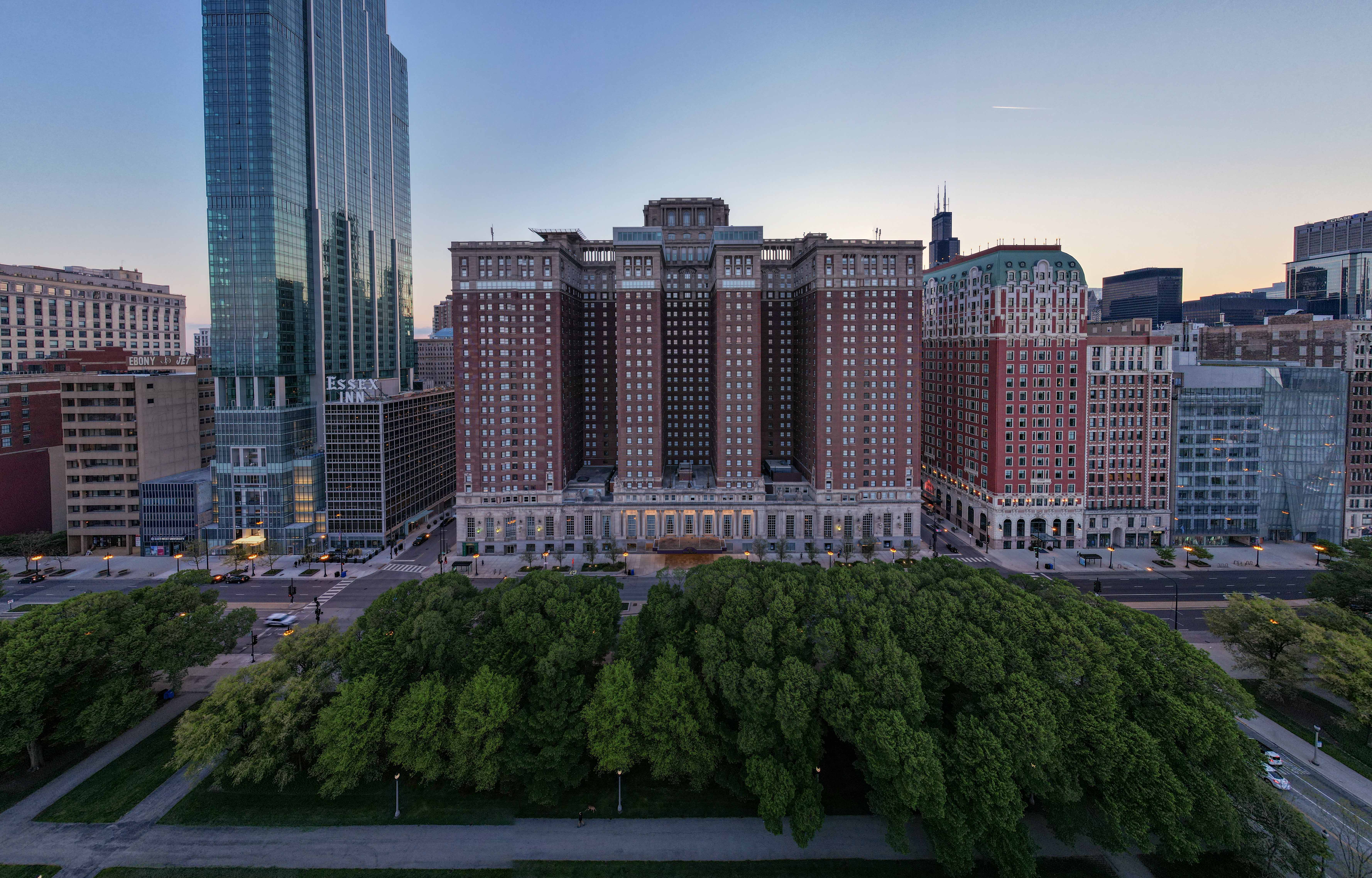 Large historic building of the Hilton Chicago flanked by modern skyscrapers, with a dense green park in the foreground at dusk.