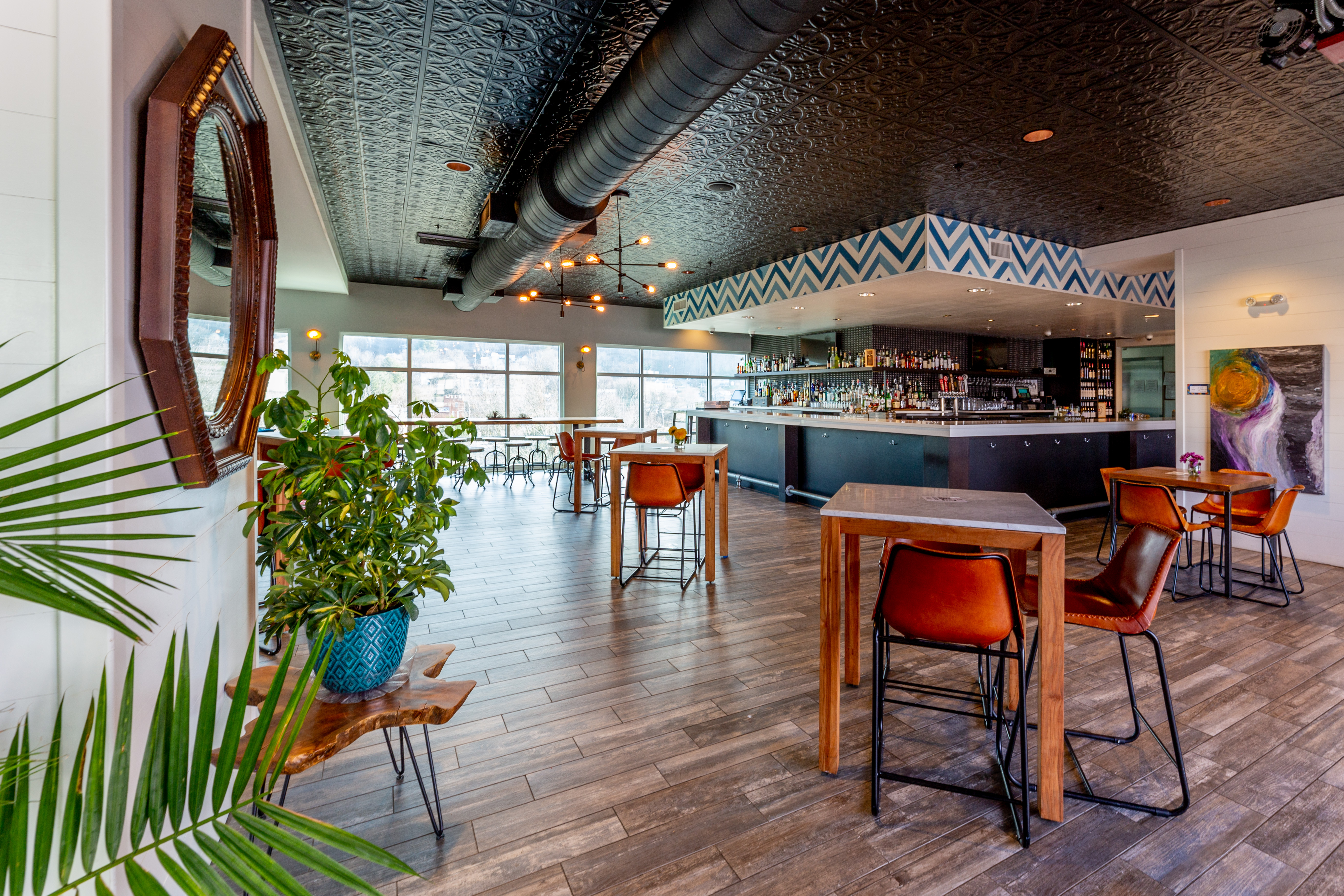 Modern bar interior with wooden tables, orange chairs, large windows, and a decorative black ceiling with exposed ductwork.