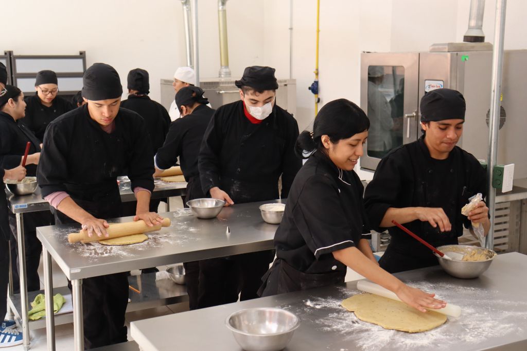 Group at International Youth Foundation in black uniforms rolling dough and preparing food in a professional kitchen.