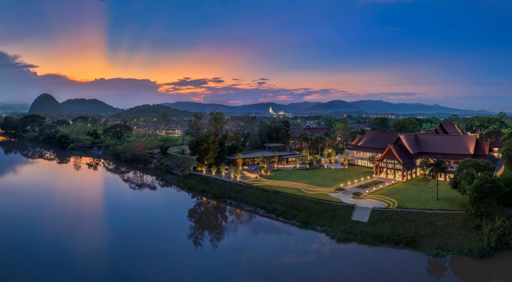 Riverfront Kahavadi Chiang Rai, Curio Collection by Hilton hotel illuminated at dusk, surrounded by trees and mountains under a colorful sunset sky.