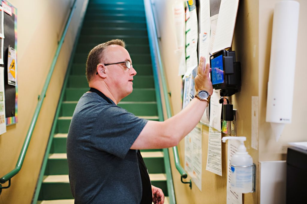 Man at National Down Syndrome Society with glasses and a hearing aid using a wall-mounted touchscreen device near a staircase.