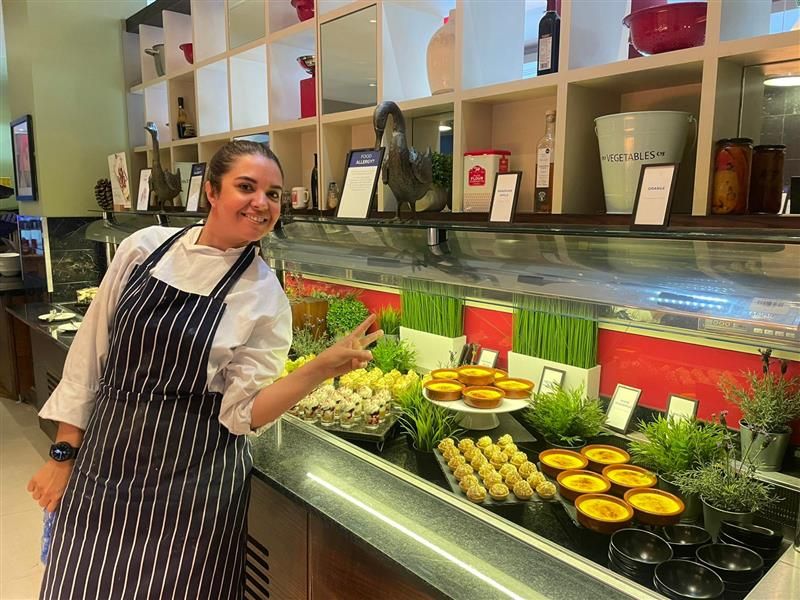 Person in a striped apron smiling and posing with a peace sign next to a dessert buffet with various pastries and greenery at Only a Pavement Away.