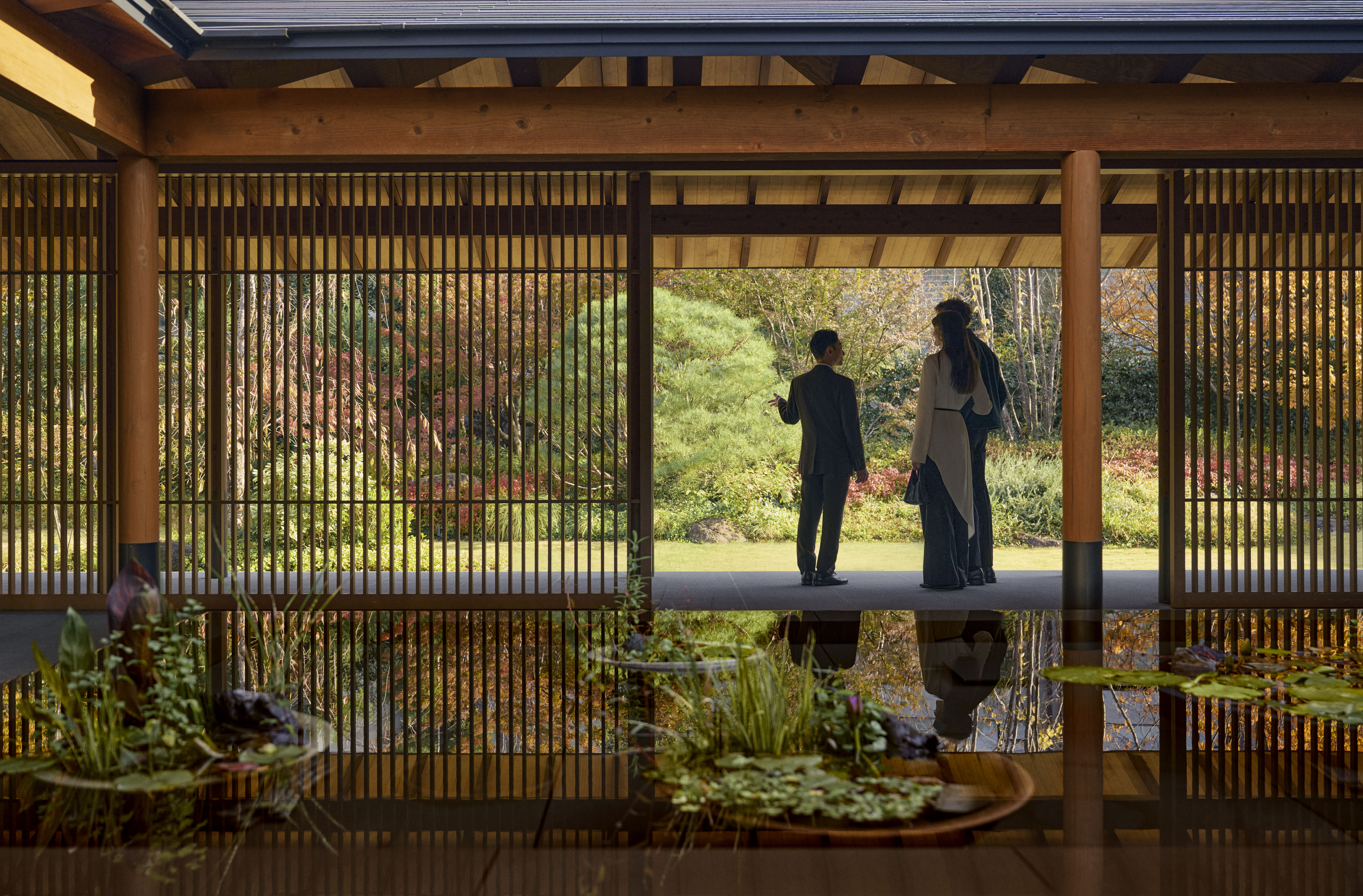 Three people standing and talking in a wooden pavilion with vertical slats, overlooking a garden at ROKU KYOTO, LXR Hotels & Resorts.