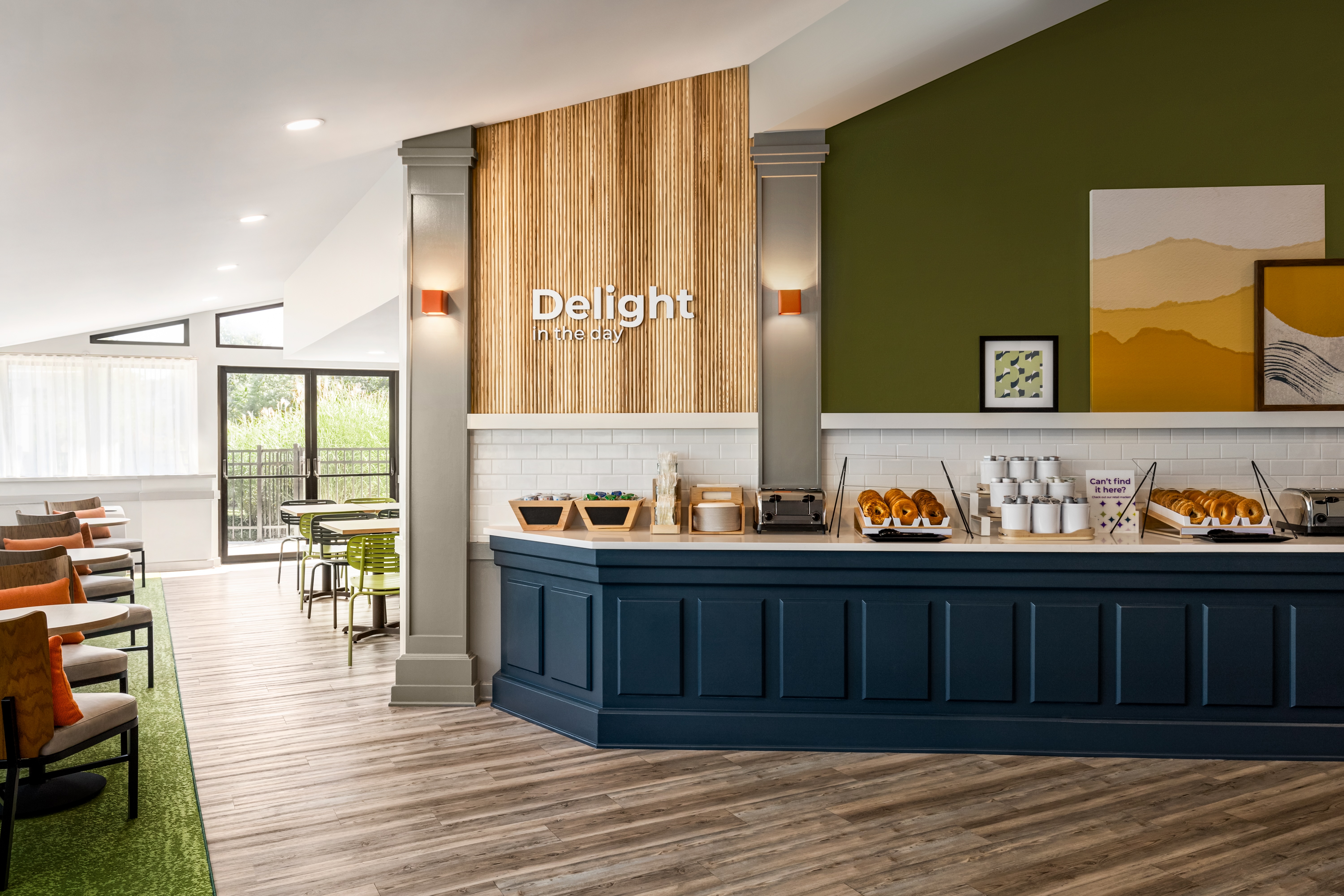 Modern breakfast area with a blue counter, bagels, condiments, and seating with orange pillows and green chairs.