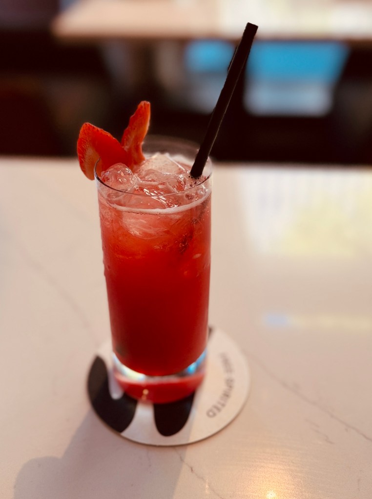 Red iced drink garnished with strawberry slices and a black straw in a tall glass on a coaster.