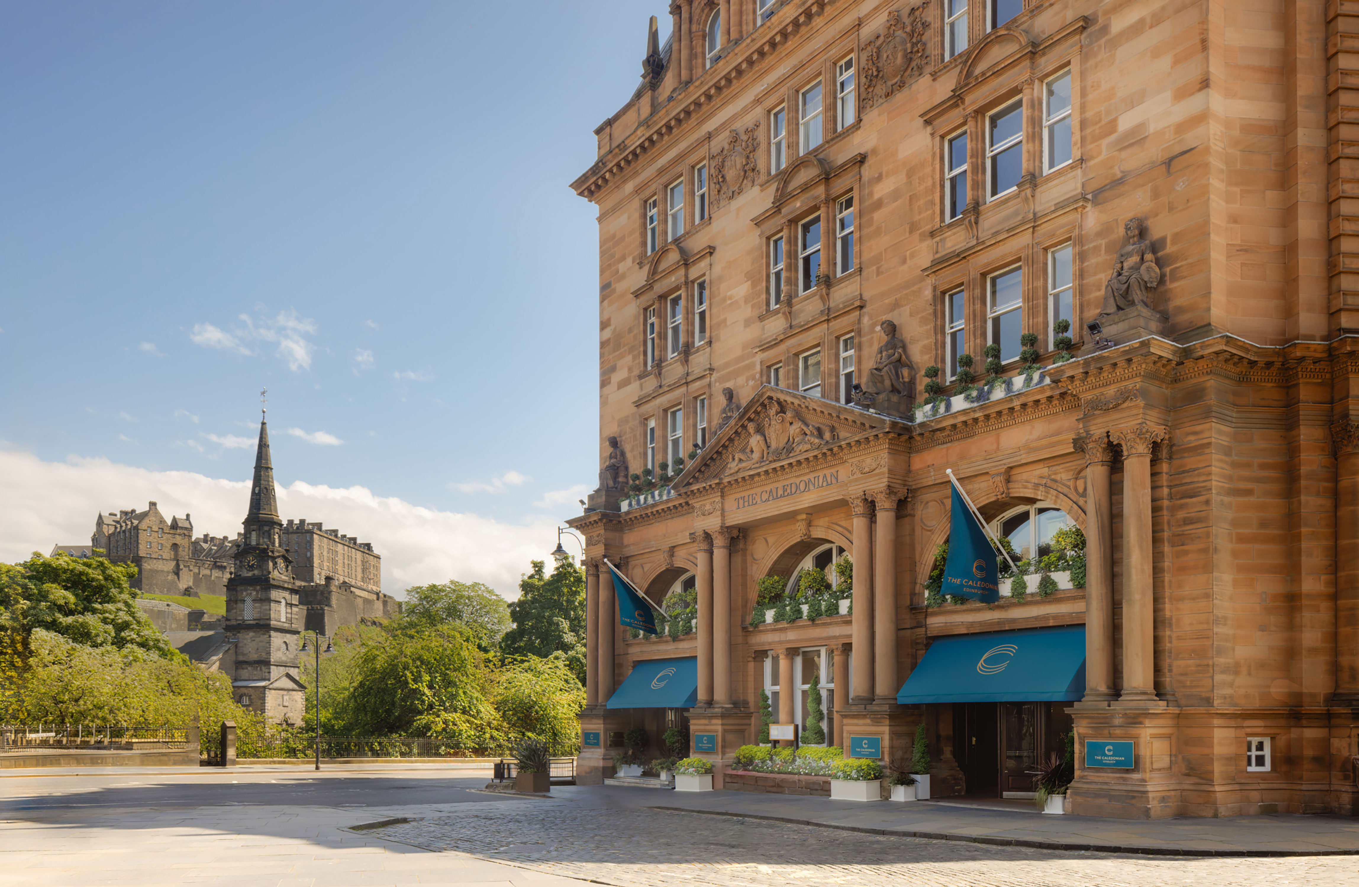 Historic sandstone building of The Caledonian Edinburgh, Curio Collection by Hilton with blue flags and awnings, with a church spire and castle in the background.
