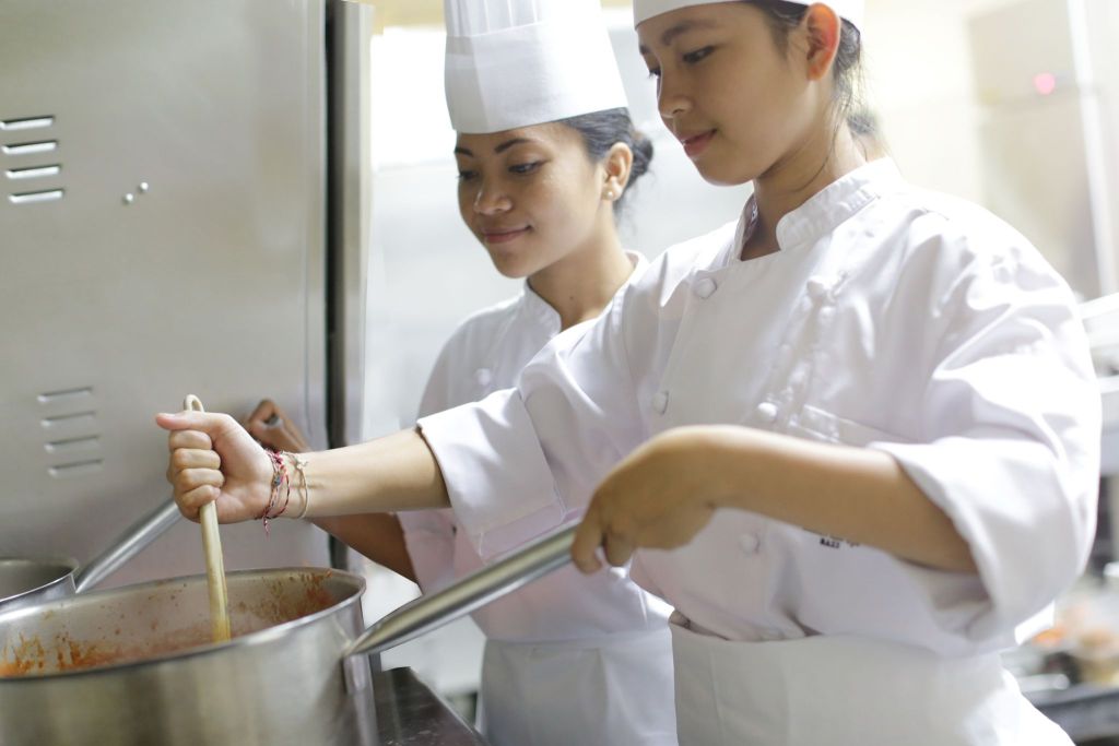 Two chefs in white uniforms and hats cooking and stirring food in a large pot in a professional kitchen at The R.O.L.E. Foundation (Rural Outreach and Livelihood Education).
