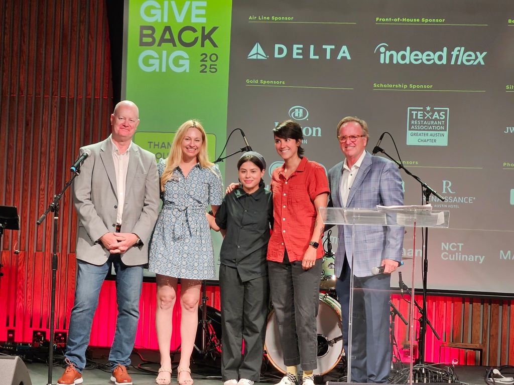 Five people stand on stage at Travis Early High School Hospitality College in front of a screen displaying sponsor logos for the "Give Back Gig 2025" event.