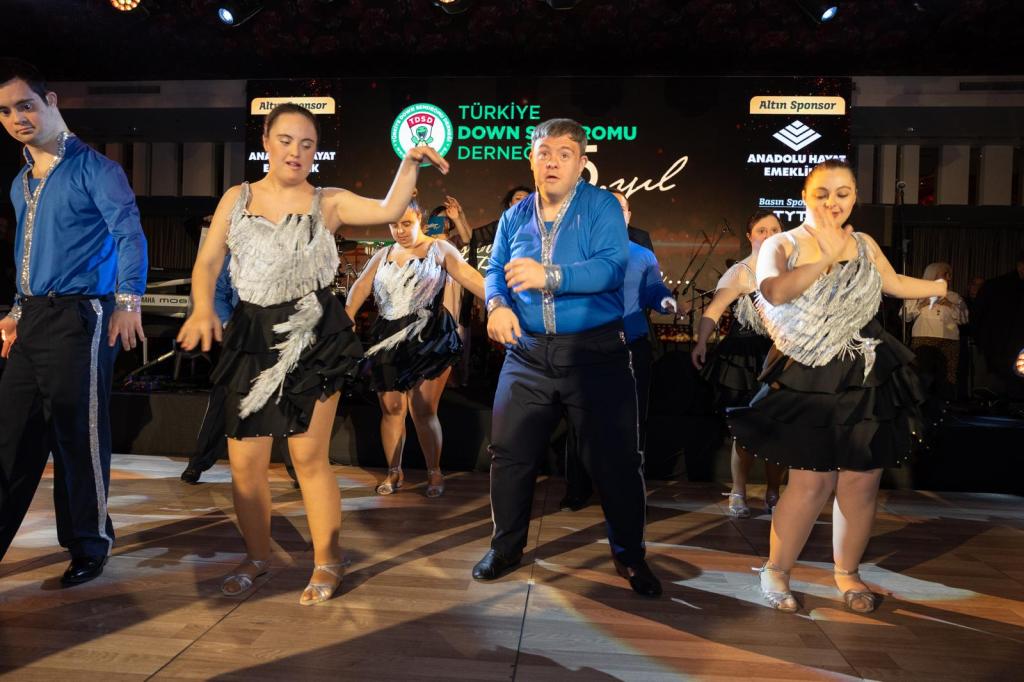 A group of dancers at Türkiye Down Syndrome Association with Down syndrome performing on stage wearing coordinated blue and silver costumes.