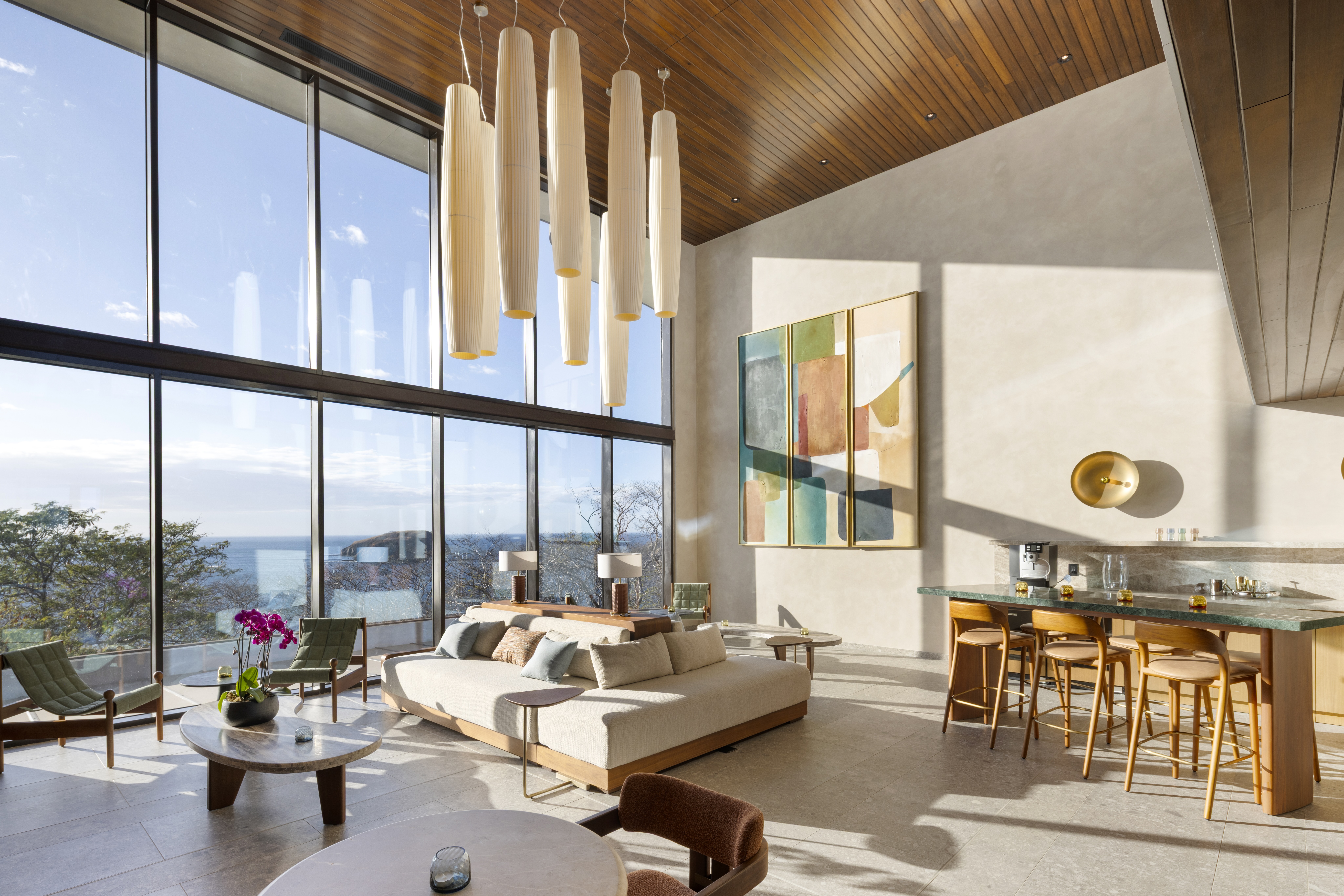 Waldorf Astoria Costa Rica Punta Cacique - Residences - Owners Lounge - Spacious modern living room with large floor-to-ceiling windows overlooking the ocean, featuring a beige sectional sofa, wooden chairs, and a kitchen island with bar stools.