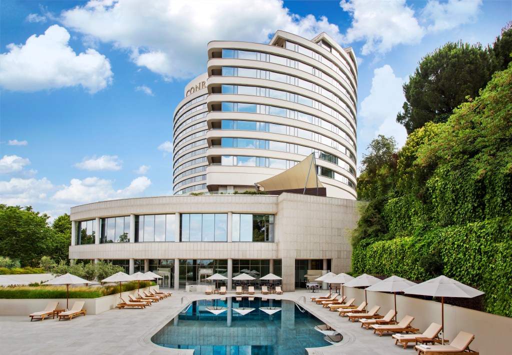 Modern curved Conrad Istanbul Bosphorus building with a pool surrounded by lounge chairs and umbrellas, next to a green ivy-covered wall.