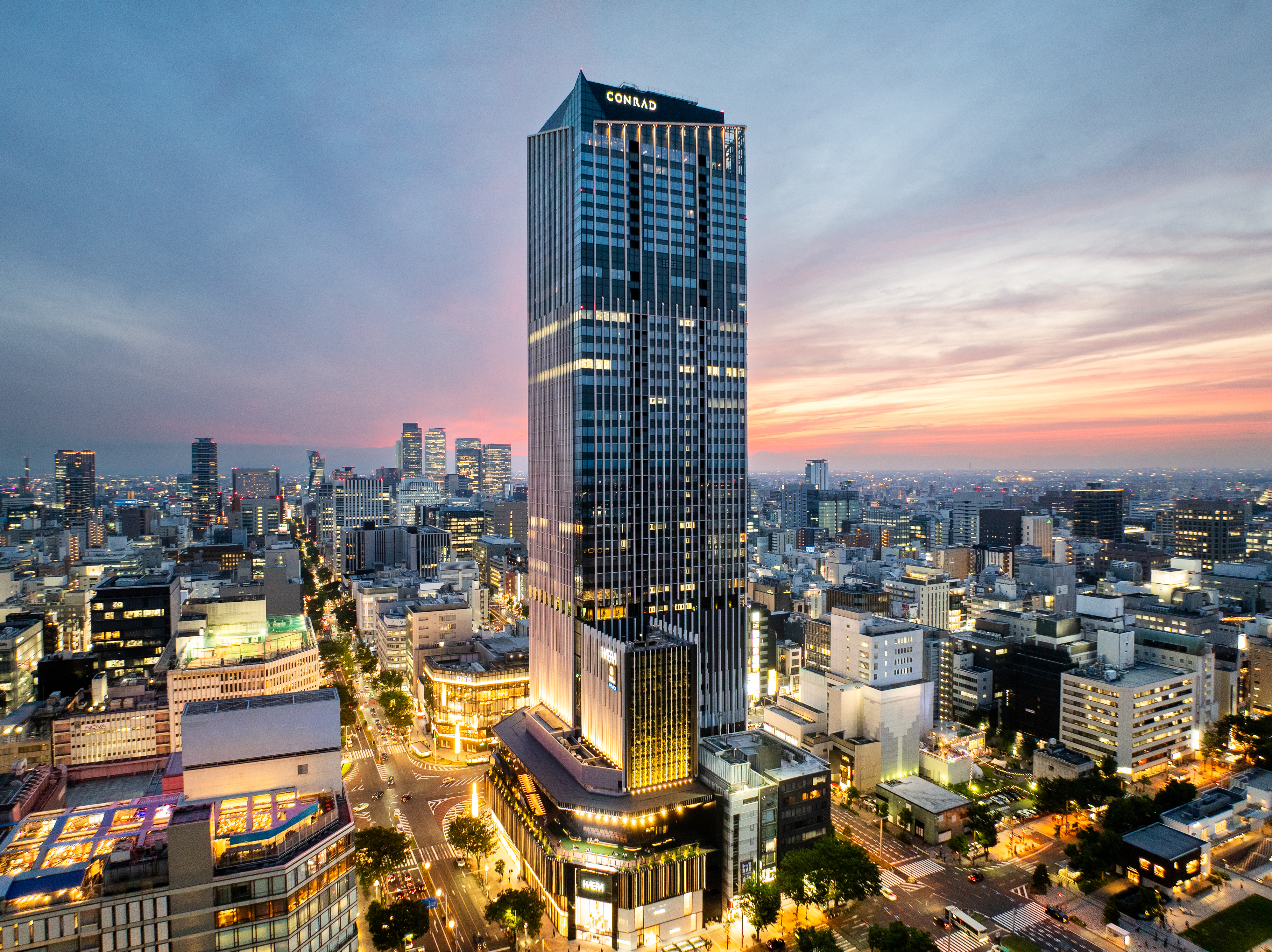 Tall modern skyscraper with "CONRAD" sign at the top, surrounded by city buildings at sunset with a colorful sky.