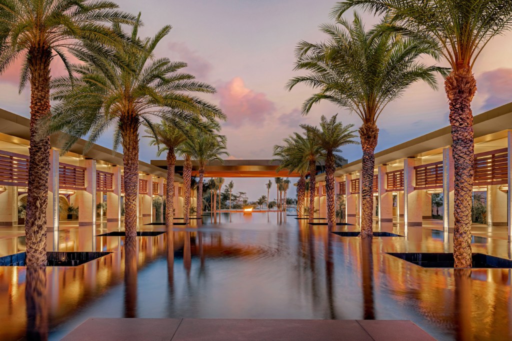 Conrad Tulum - palm trees lining a reflective water feature at sunset with a modern architectural structure in the background.