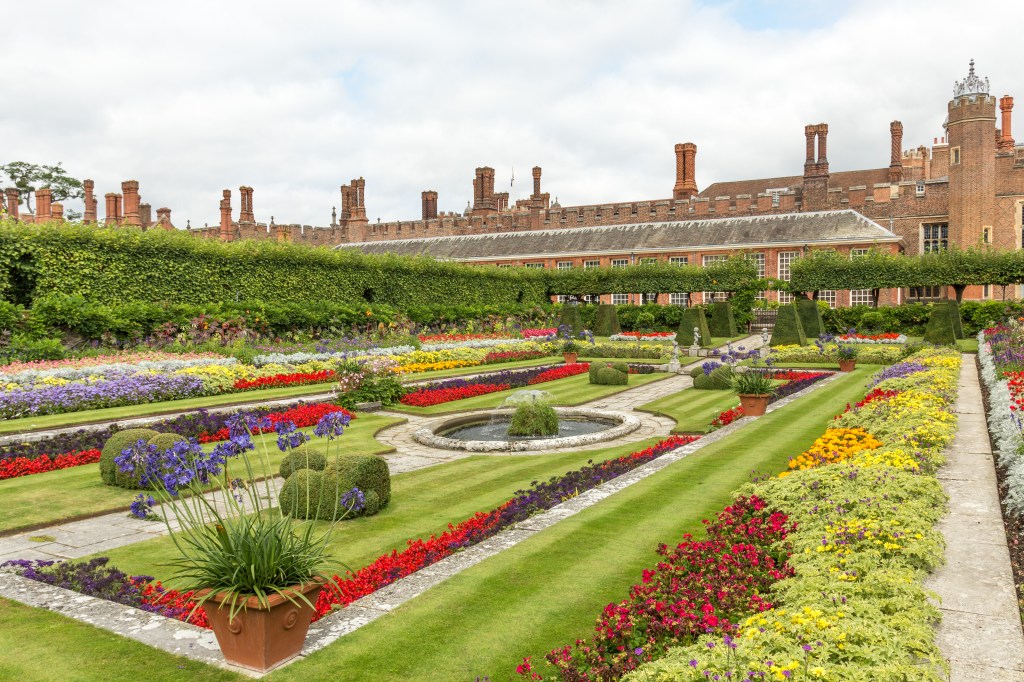 Formal garden with colorful flower beds, trimmed hedges, a central fountain, and a historic brick building in the background.