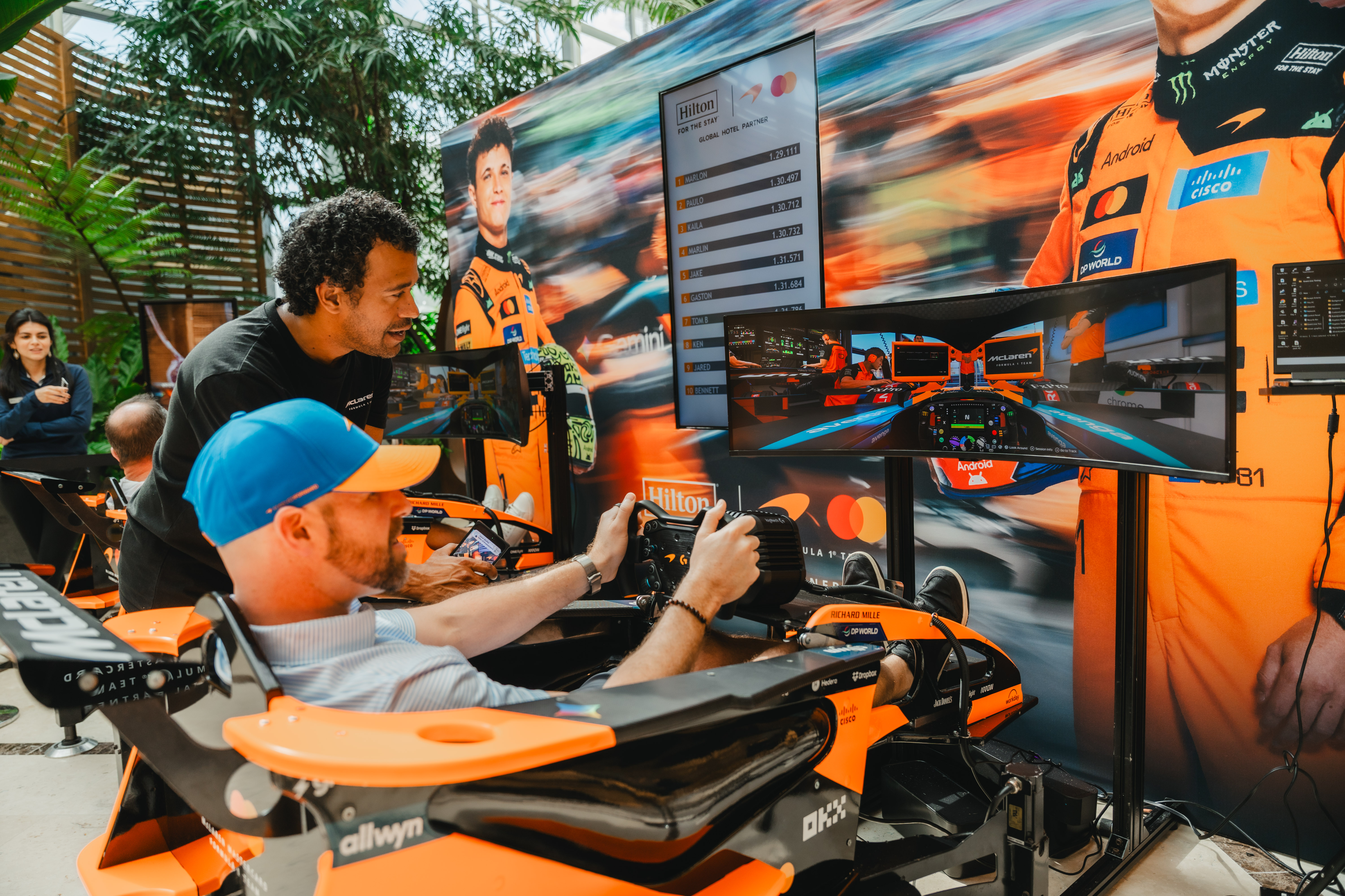 Man in blue cap using a Formula 1 racing simulator at Hilton x McLaren Racing - Miami assisted by another man wearing a black shirt.