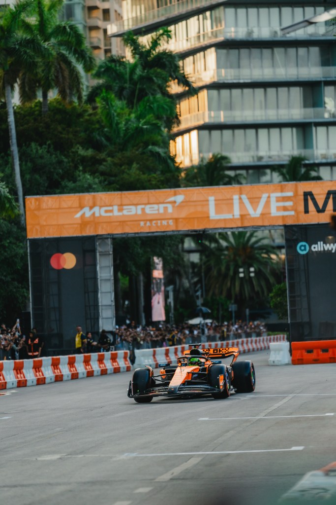 Formula 1 car racing at a Hilton x McLaren Racing experience in Miami city street circuit with McLaren Racing banners and spectators behind barriers.