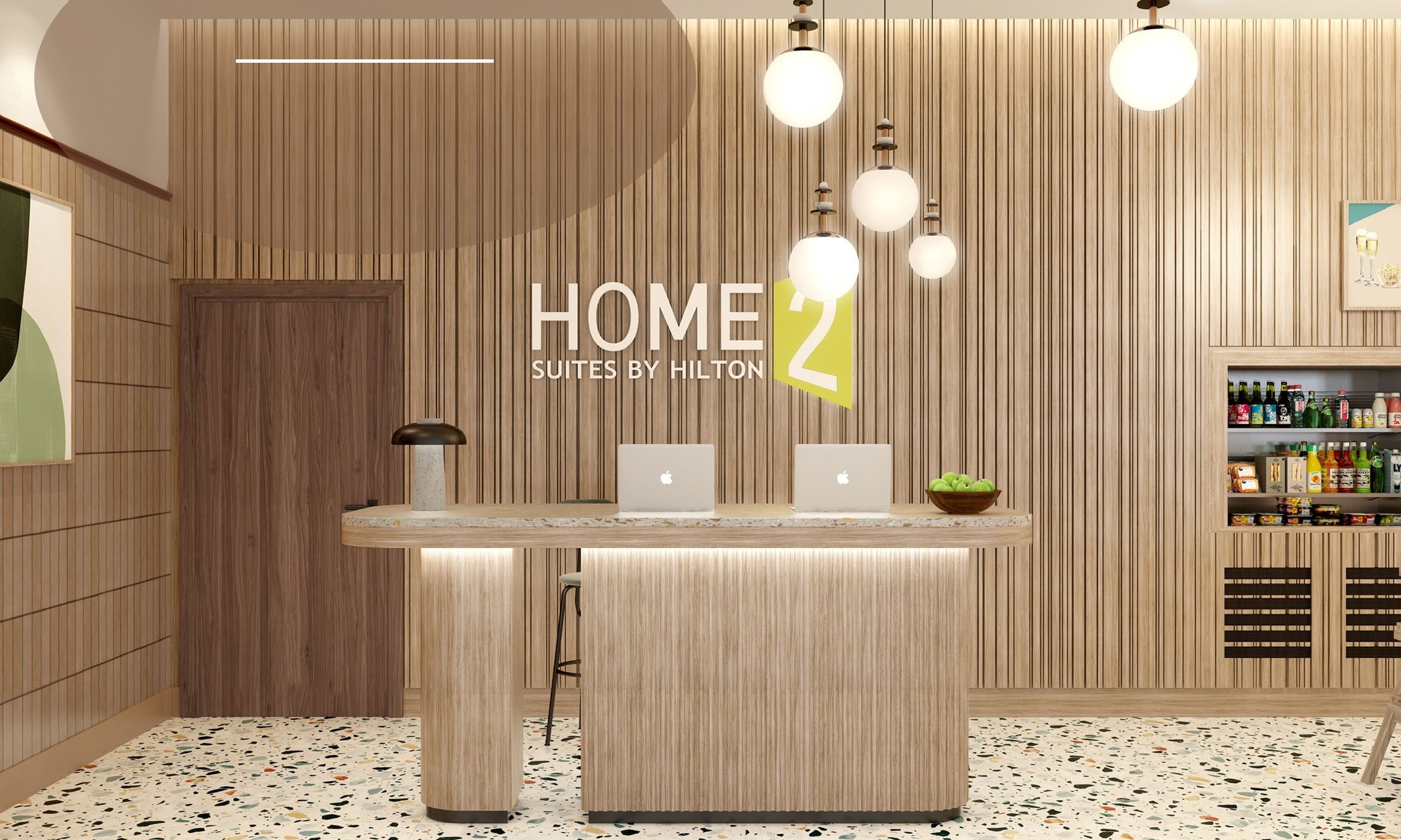 Reception area of Home2 Suites by Hilton with a wooden desk, two laptops, hanging globe lights, and a small snack shelf on the right.