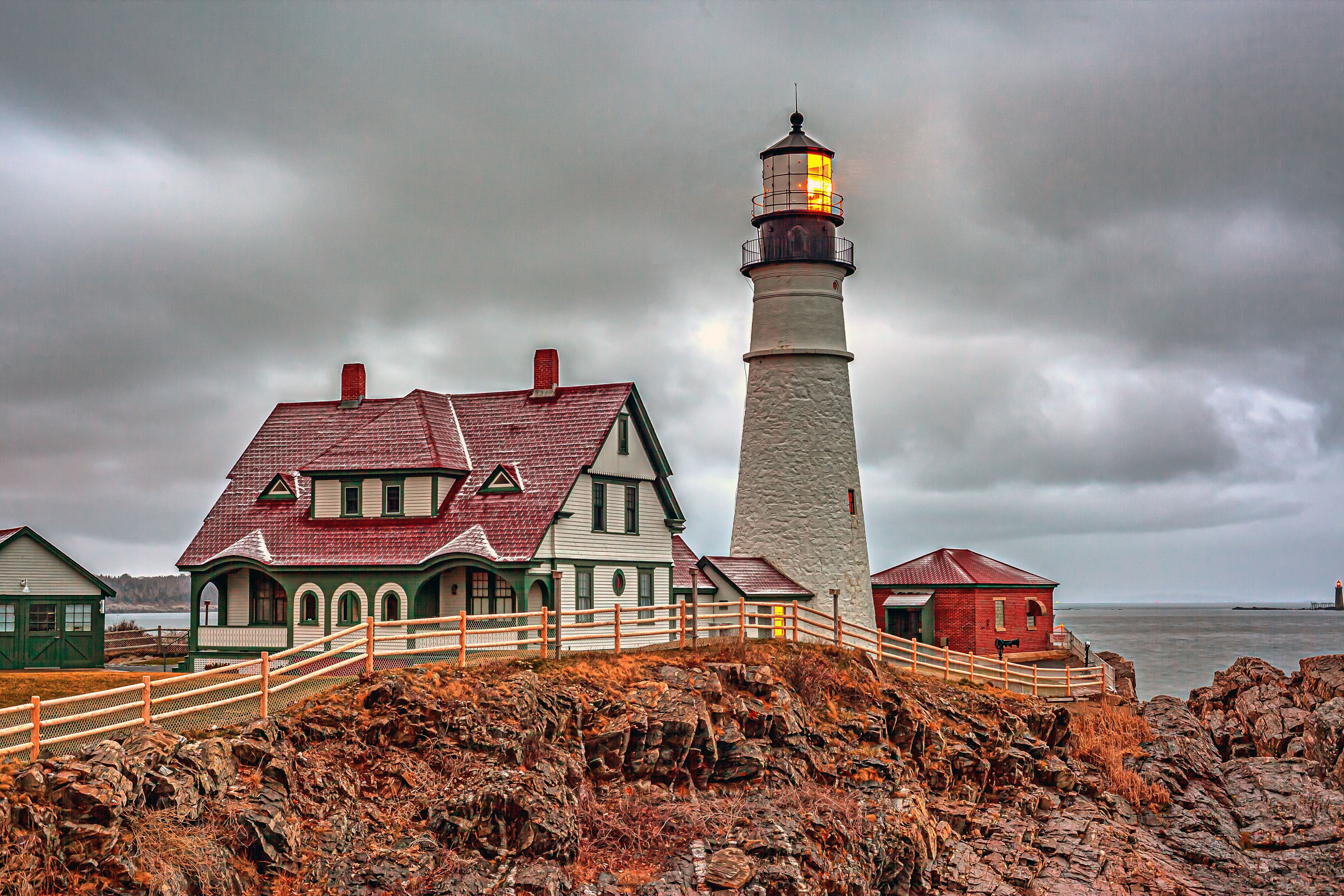 Lighthouse with a glowing light next to a house with a red roof on rocky coastline under a cloudy sky.