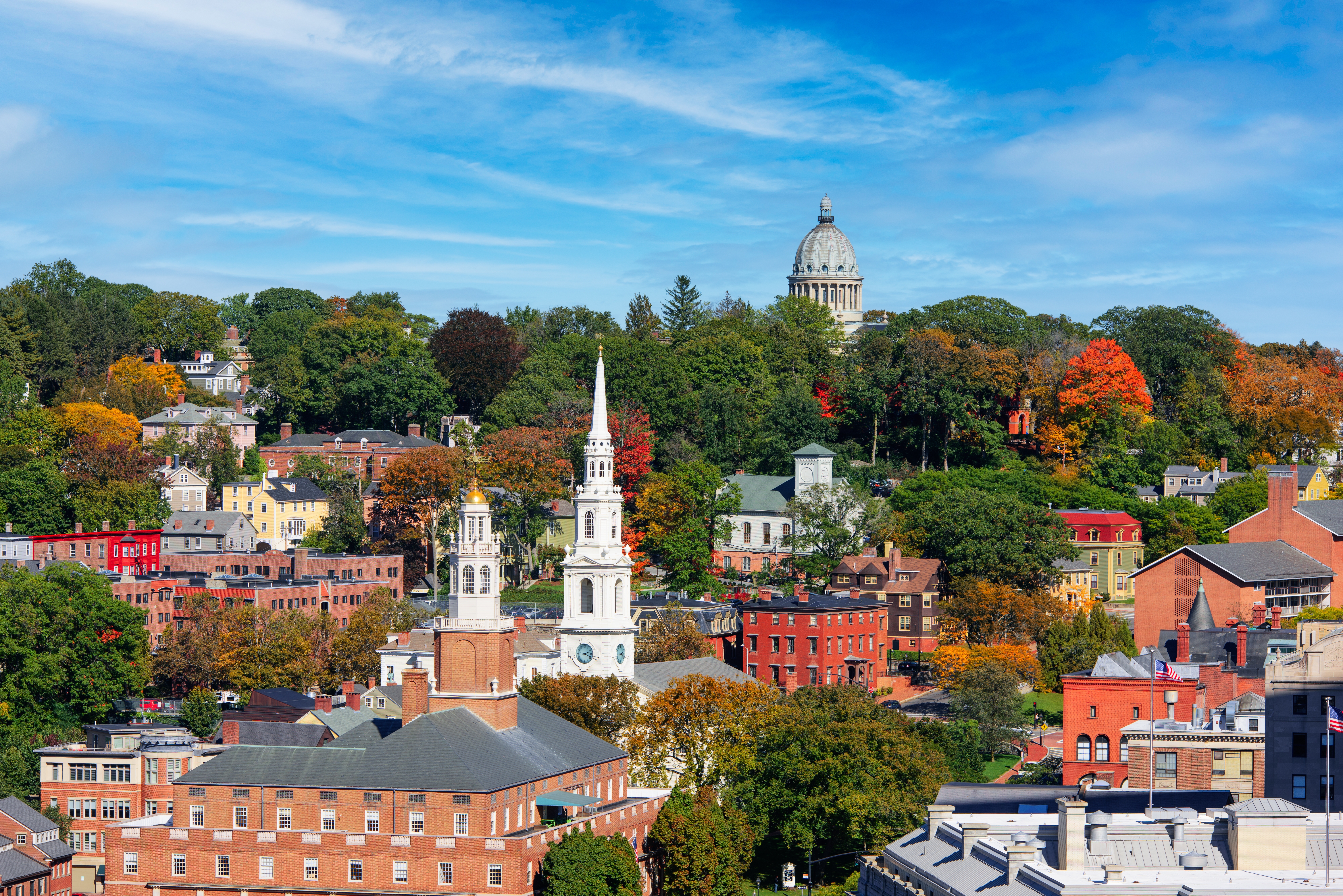 Colorful autumn trees surround historic brick buildings and church steeples under a bright blue sky.