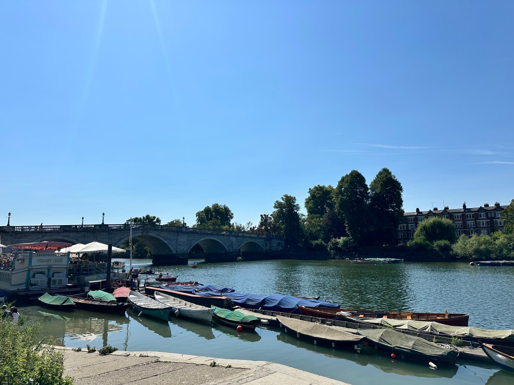 River with moored boats, a stone bridge in the background, and trees and buildings along the riverbank under a clear blue sky.