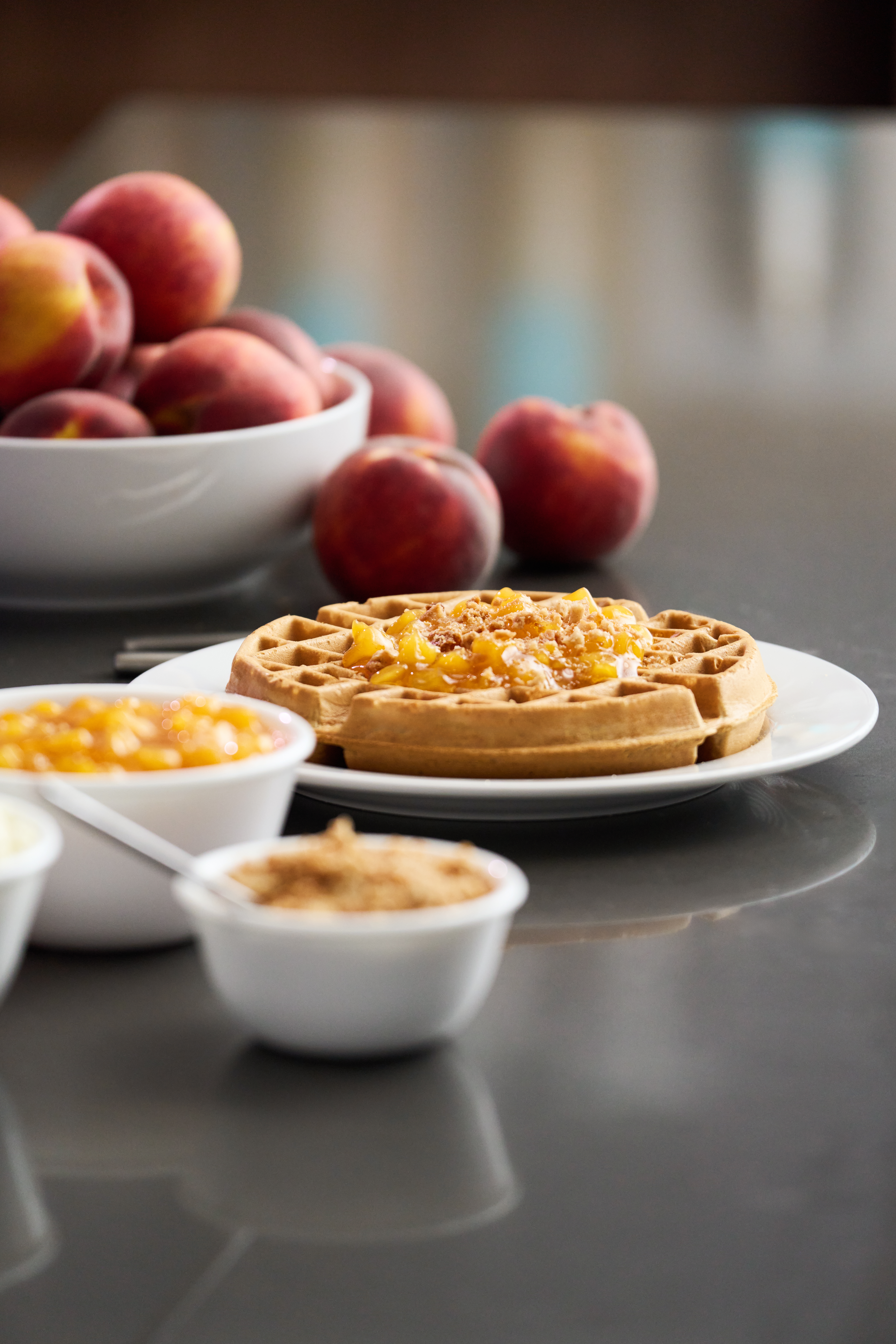 Waffle topped with fruit compote on a white plate, with bowls of toppings and a bowl of peaches in the background.