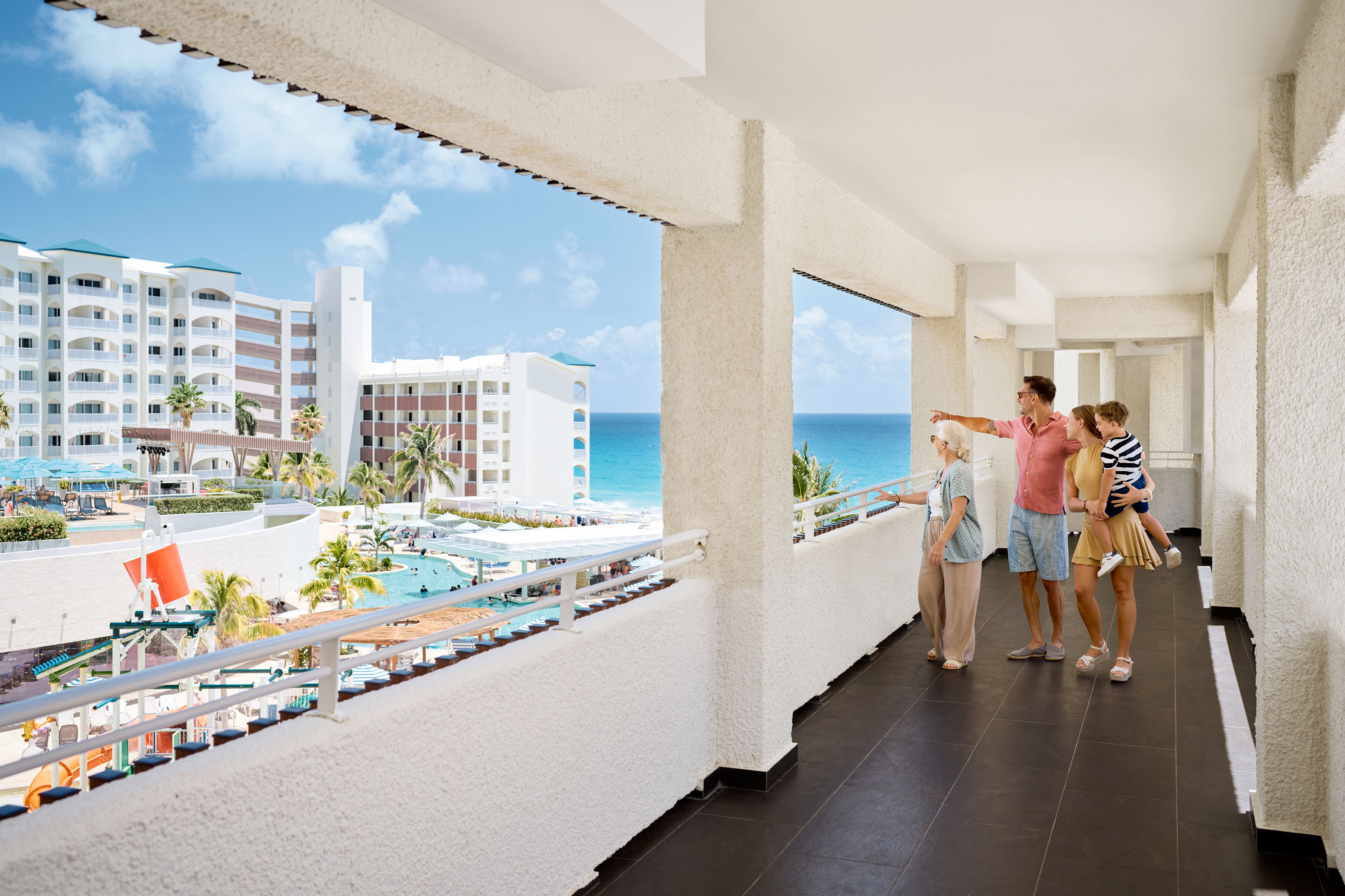 Family of four on a hotel balcony overlooking a pool, palm trees, and the ocean on a sunny day at Hilton Cancun Mar Caribe All-Inclusive Resort.