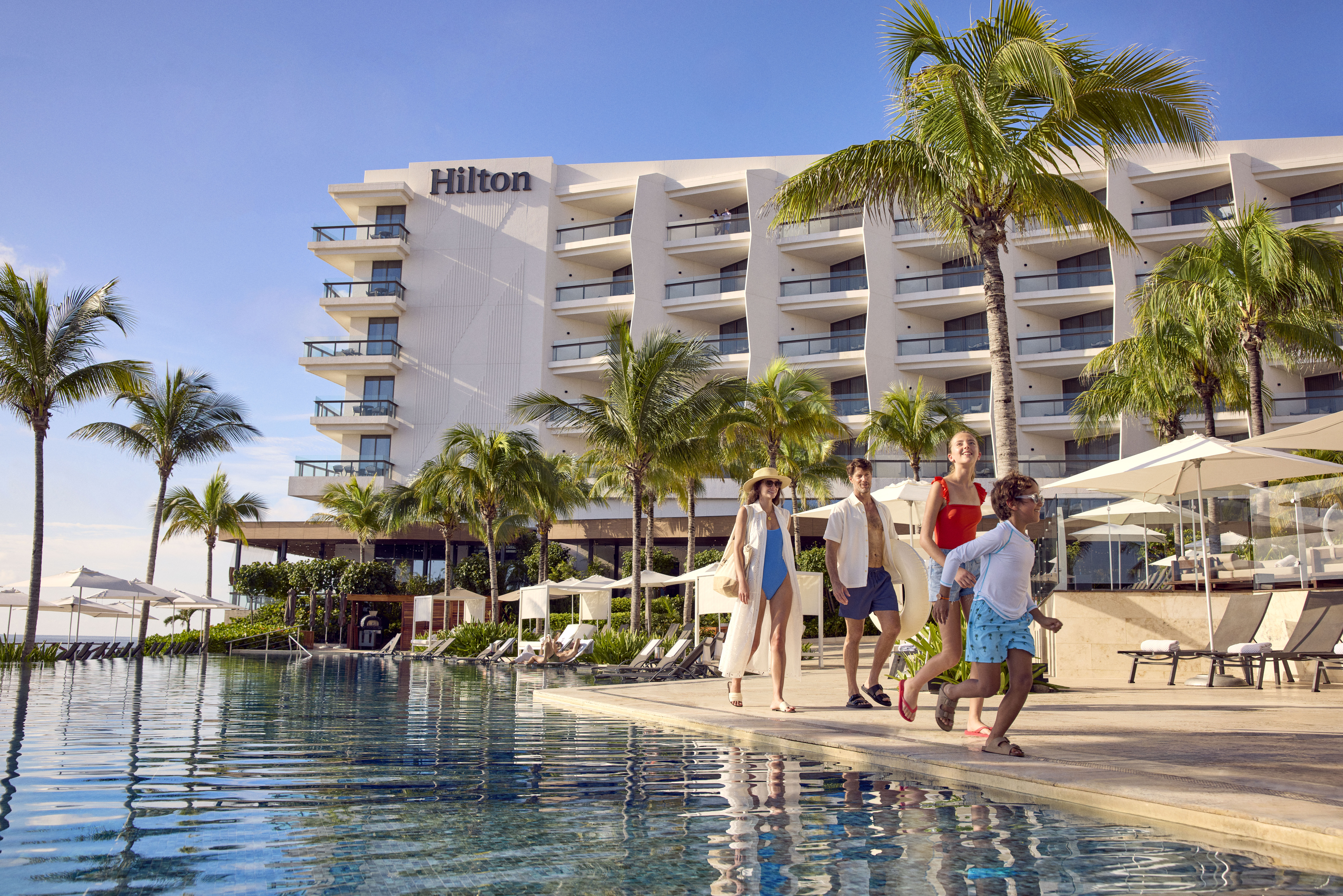 Family walking by the pool at Hilton Cancun, an All-Inclusive Resort with palm trees and lounge chairs under umbrellas.